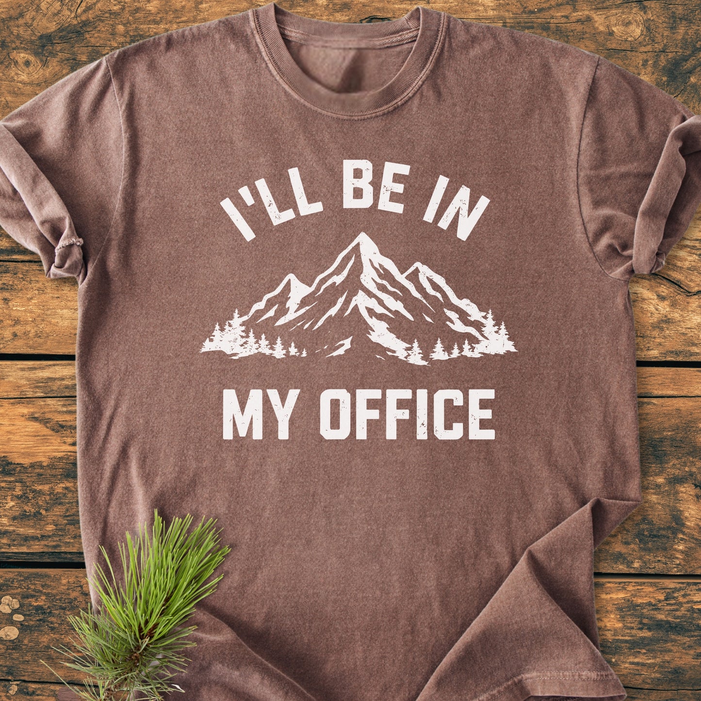 I'll Be In My Office - Vintage Wash Tee featuring a mountain and pine graphic on a brown, crew-neck T-shirt, laid flat on wood with a pine sprig accent.