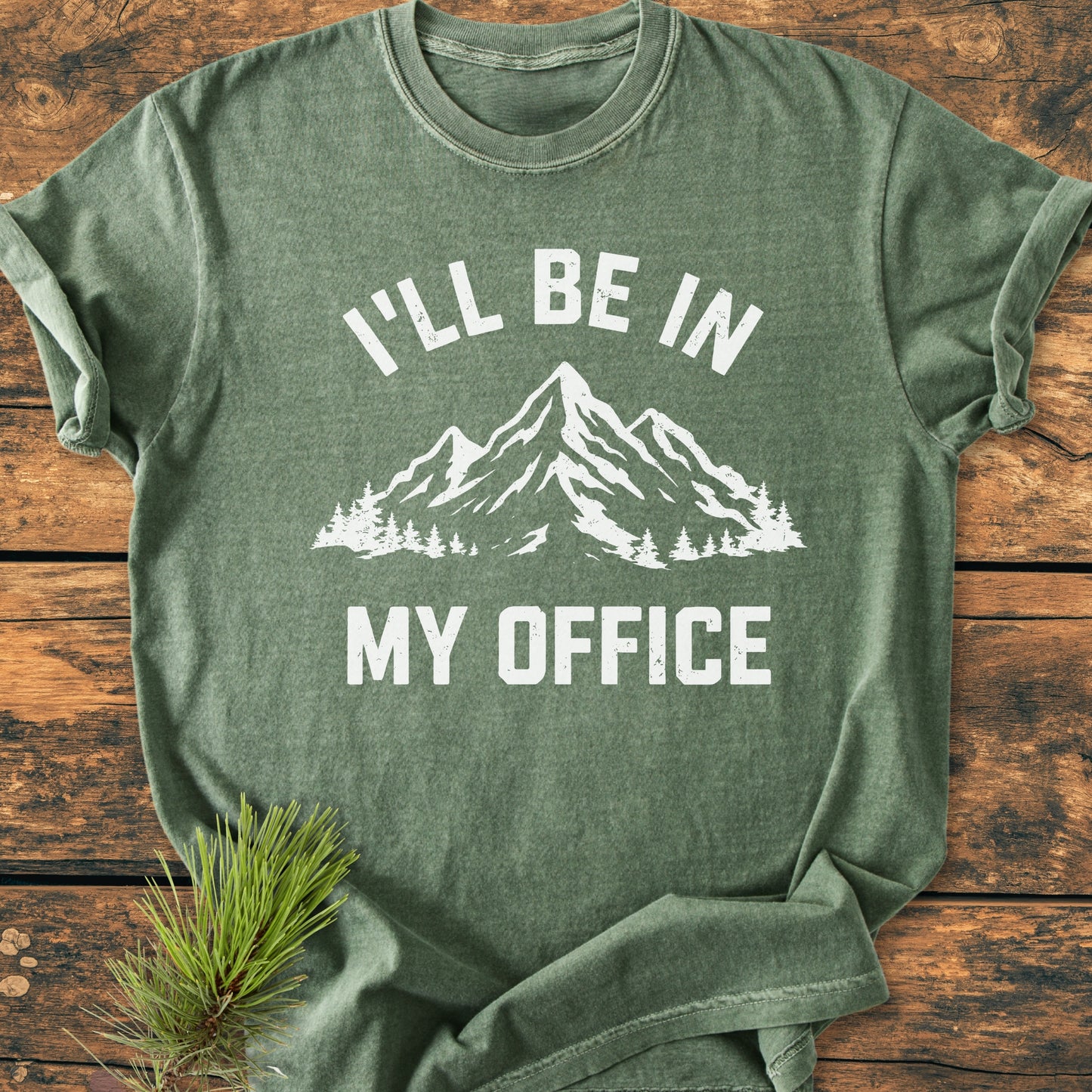 I'll Be In My Office - Vintage Wash Tee featuring a white mountain and tree graphic on a forest-green background, laid on wood with a pine sprig, showcasing comfort and durability.