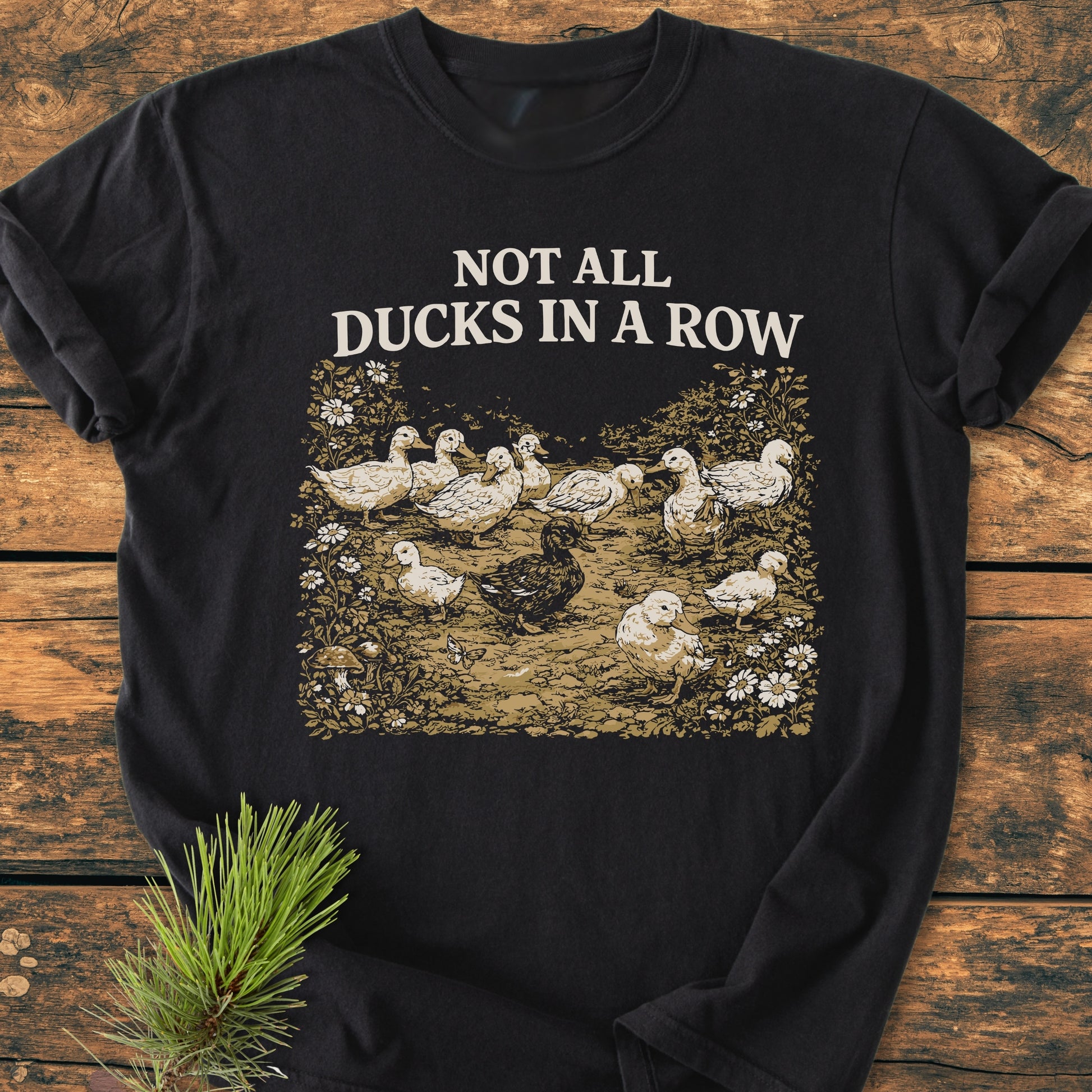 Not All Ducks In A Row - Vintage Wash Tee on wood, features playful duck illustration with a standout darker duck. Includes a pine sprig beside it. Comfortable, relaxed fit, and durable design.