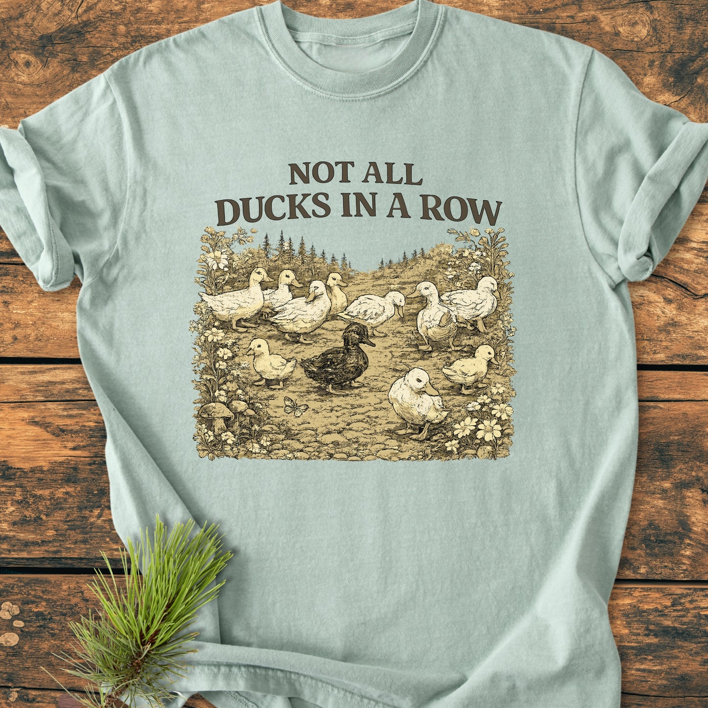Not All Ducks In A Row - Vintage Wash Tee features a vintage duck illustration, one dark among light, with foliage. Laid out on wood with a pine sprig accent, highlighting its relaxed, durable design.