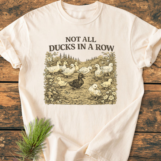 Not All Ducks In A Row - Vintage Wash Tee with scattered duck illustration and floral accents, laid on wood surface, showcasing a relaxed fit and soft garment-dyed fabric for everyday wear.