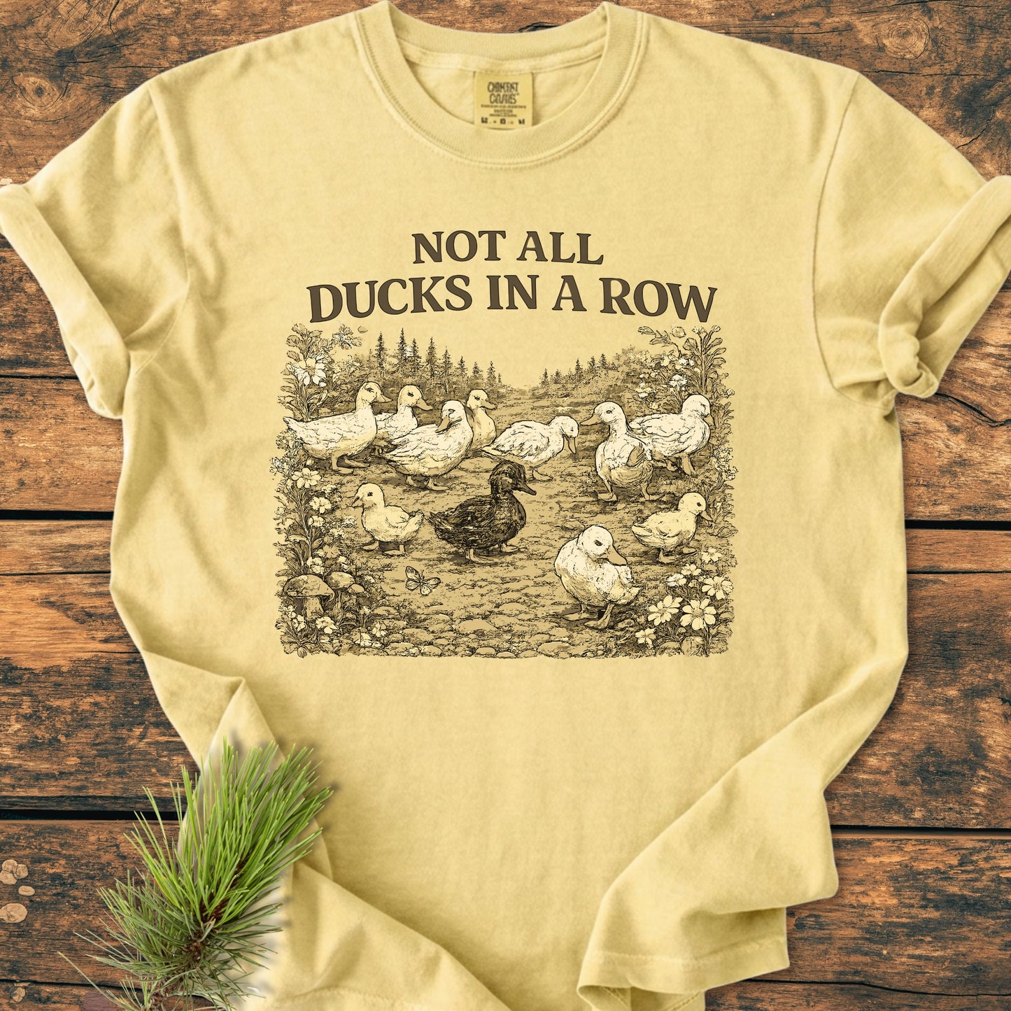 Not All Ducks In A Row - Vintage Wash Tee features a sepia-tone illustration of ducks in a clearing with foliage, on a pale yellow T-shirt, emphasizing comfort with a relaxed fit and durable design.