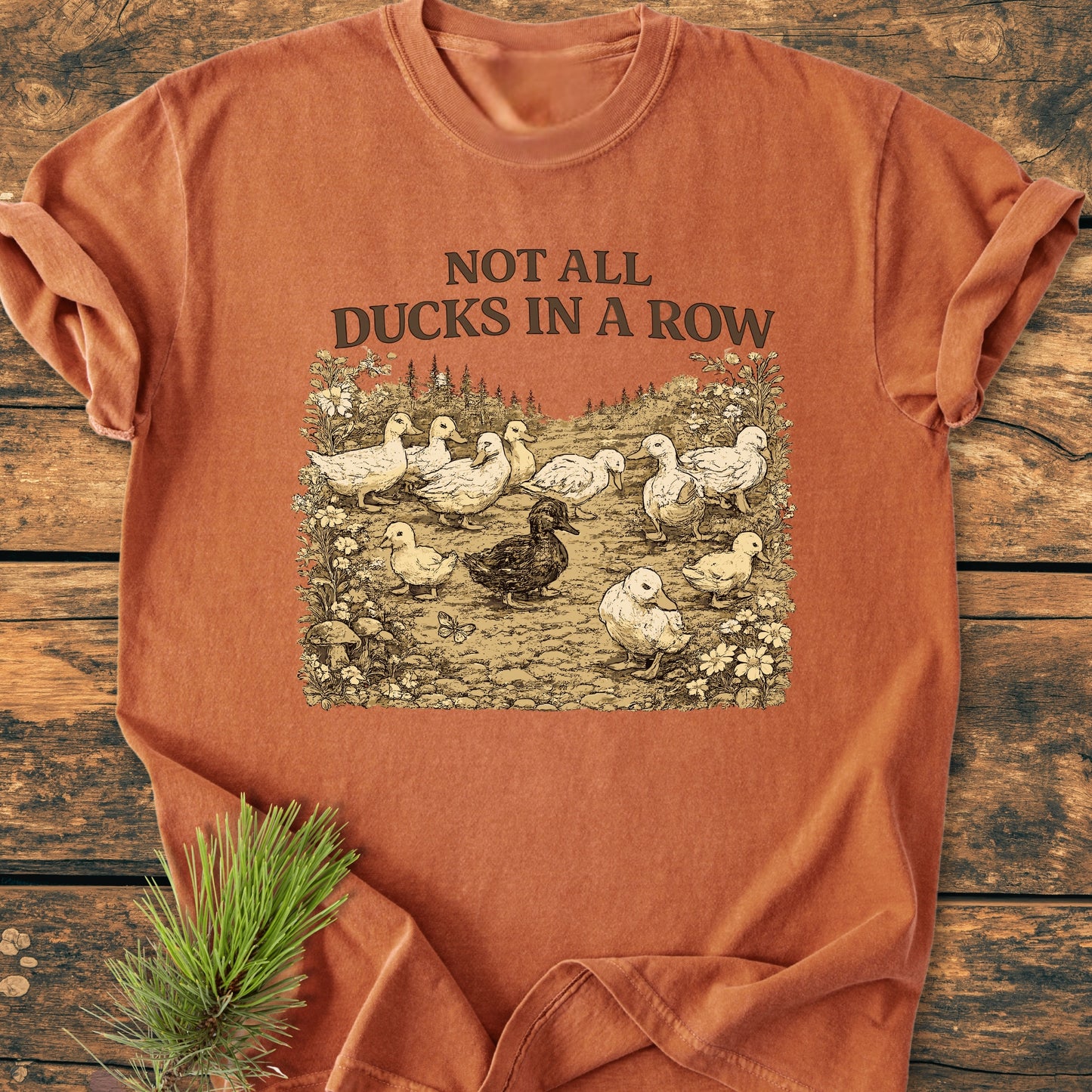 Not All Ducks In A Row - Vintage Wash Tee features a duck illustration on a rust-orange crewneck, styled on wood with a pine sprig. Comfortable, garment-dyed 100% ring-spun cotton offers a relaxed fit.