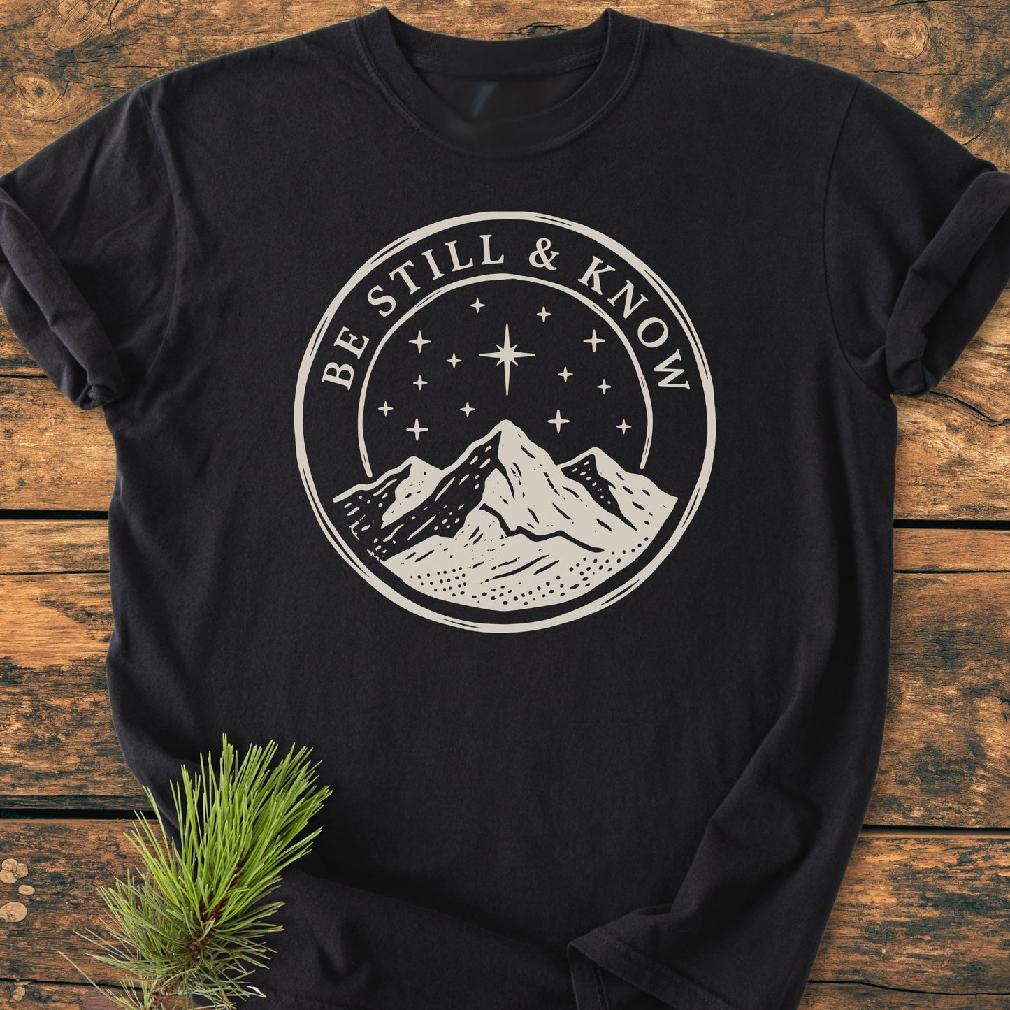Be Still And Know, Psalm 46:10 - Vintage Wash Tee featuring a mountain graphic and starry sky on soft, garment-dyed fabric, displayed on wood with rolled sleeves and pine sprig accent.