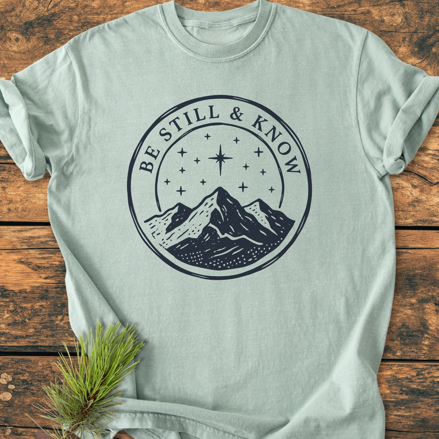 Be Still And Know, Psalm 46:10 - Vintage Wash Tee features a navy mountain graphic with stars on a pale-green shirt, laid on wood, accented by pine needles for a serene touch.