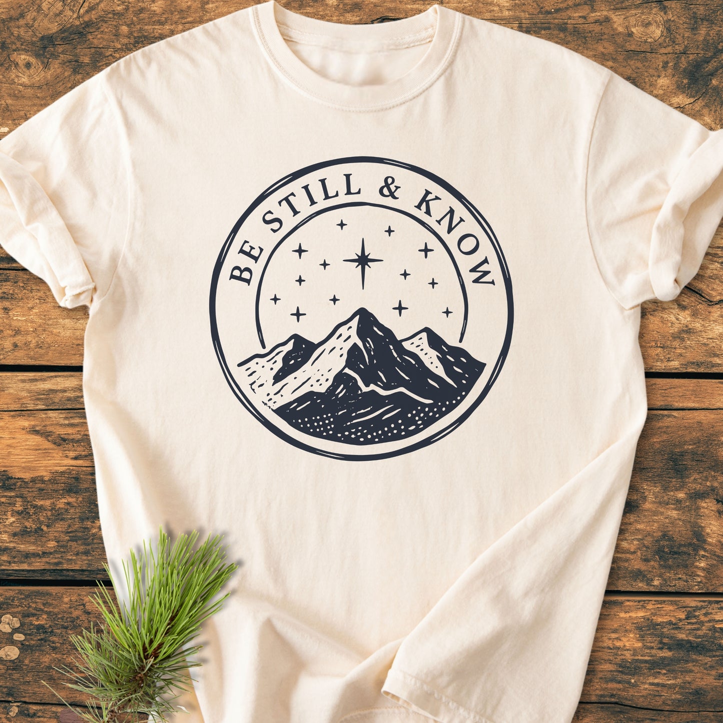 Cream-colored Be Still And Know, Psalm 46:10 - Vintage Wash Tee featuring navy-blue mountain graphic and text, laid flat on rustic wood with a pine sprig, highlighting its cozy, relaxed fit.