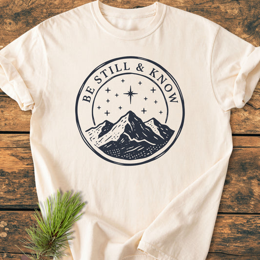 Cream-colored Be Still And Know, Psalm 46:10 - Vintage Wash Tee featuring navy-blue mountain graphic and text, laid flat on rustic wood with a pine sprig, highlighting its cozy, relaxed fit.