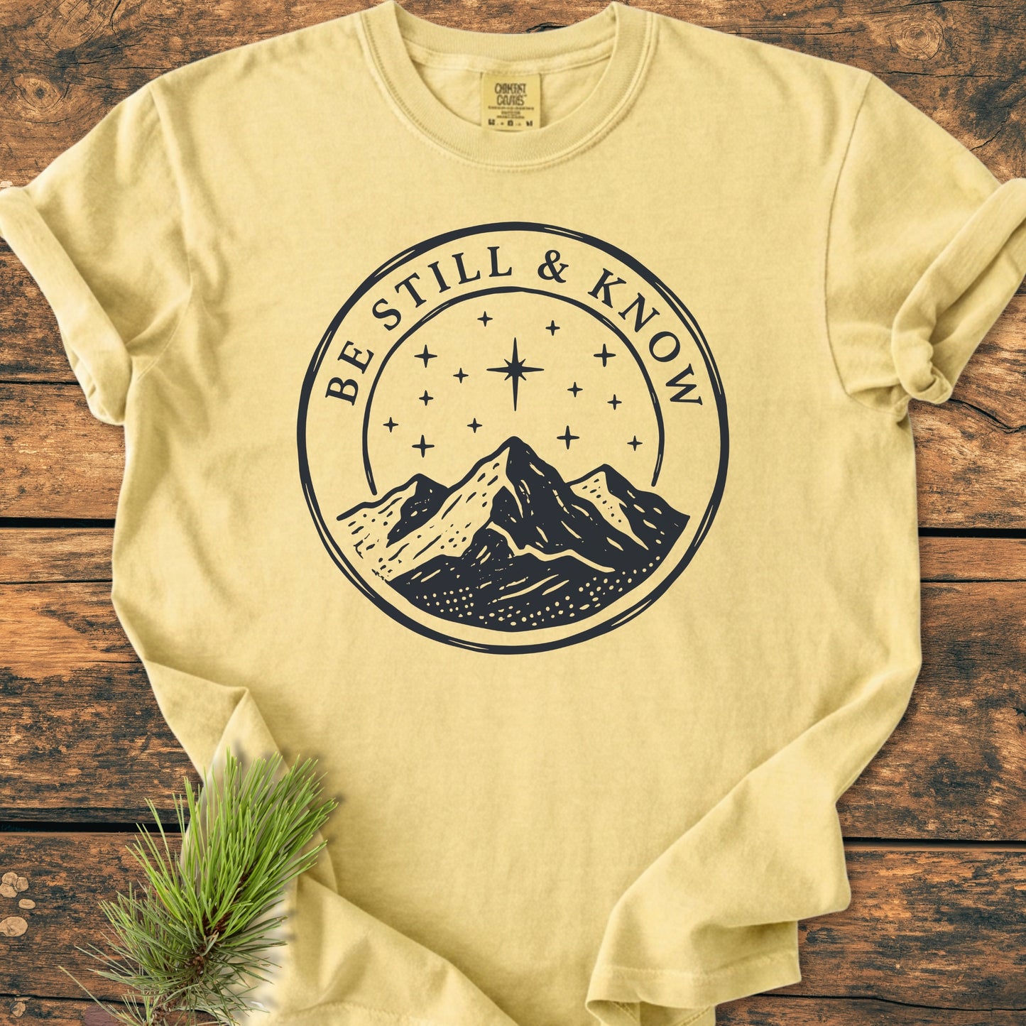 Light-yellow Be Still And Know, Psalm 46:10 - Vintage Wash Tee with a hand-drawn mountain and star graphic, laid on wood, showcasing durability and comfort of 100% ring-spun cotton.
