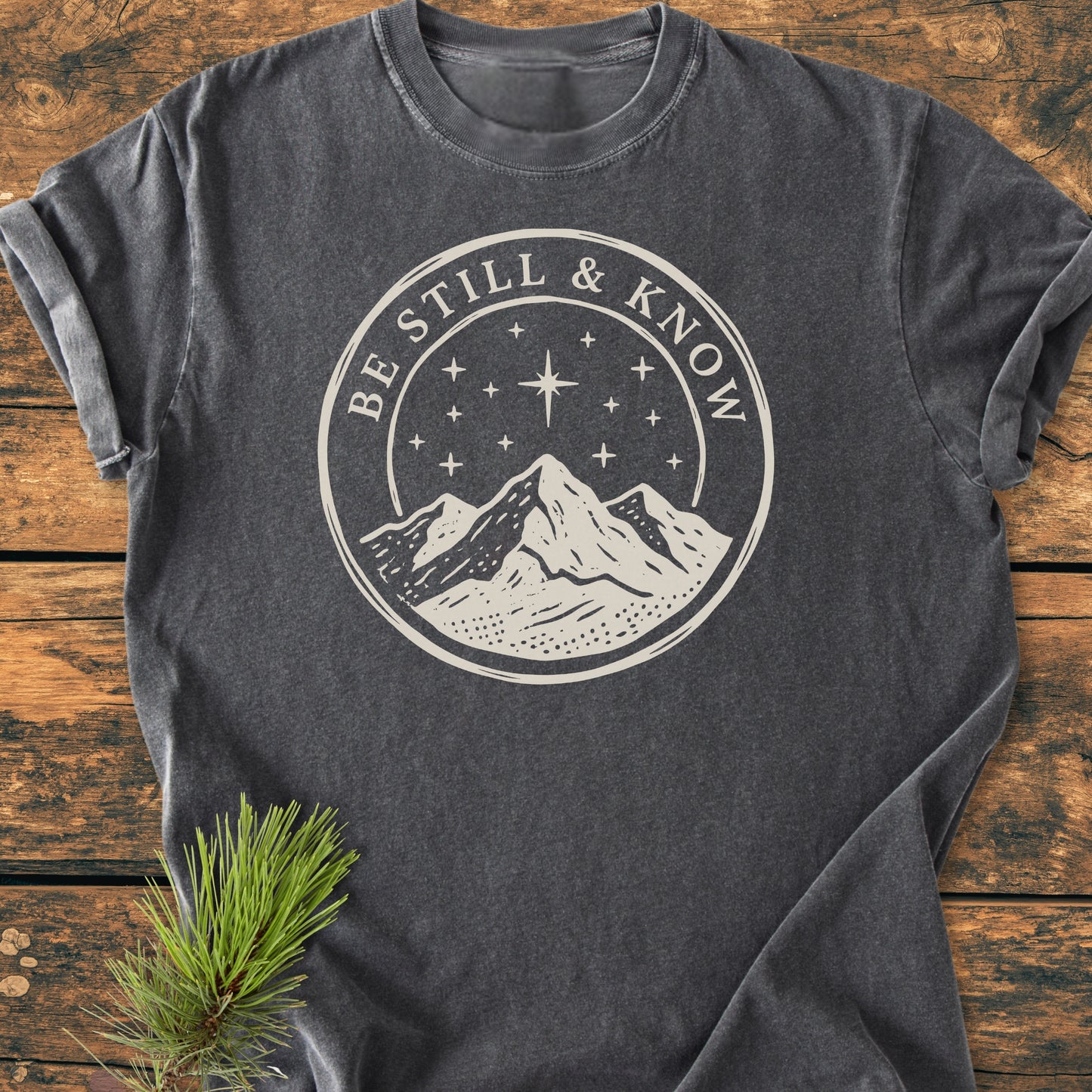 Be Still And Know, Psalm 46:10 - Vintage Wash Tee features a cream graphic of a mountain and starry sky, highlighting the phrase on a charcoal-gray Comfort Colors t-shirt laid flat.