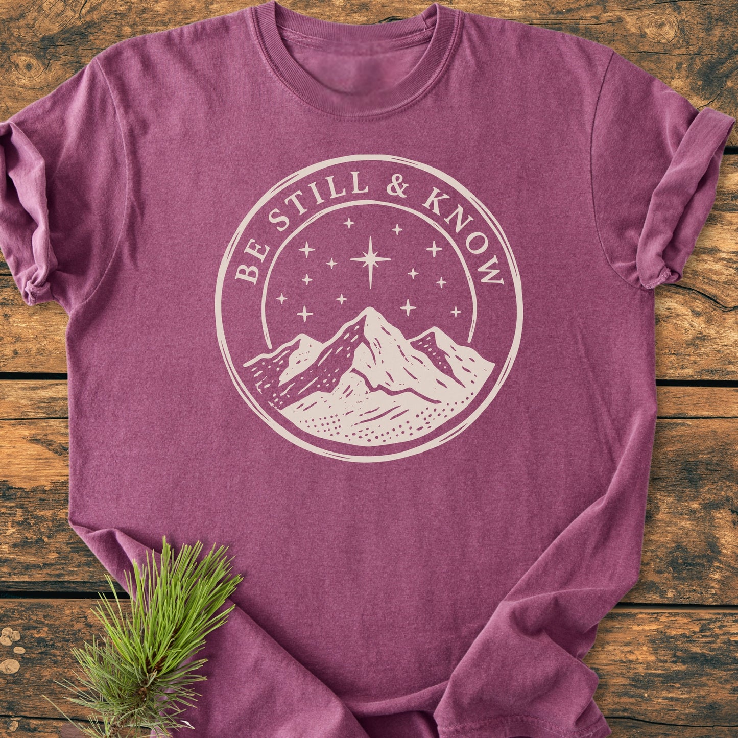 Be Still And Know, Psalm 46:10 - Vintage Wash Tee, featuring a mountain and starry sky graphic, laid on wood with pine sprig, showcases relaxed fit and durable craftsmanship in soft, garment-dyed cotton.