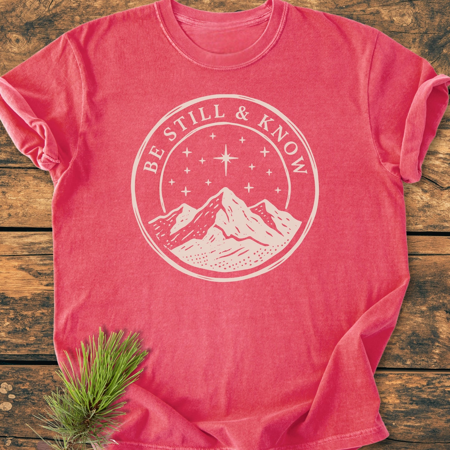 Be Still And Know, Psalm 46:10 - Vintage Wash Tee features a circular mountain graphic under a starry sky, centered on a washed-out red T-shirt, laid flat on a wood-plank background.