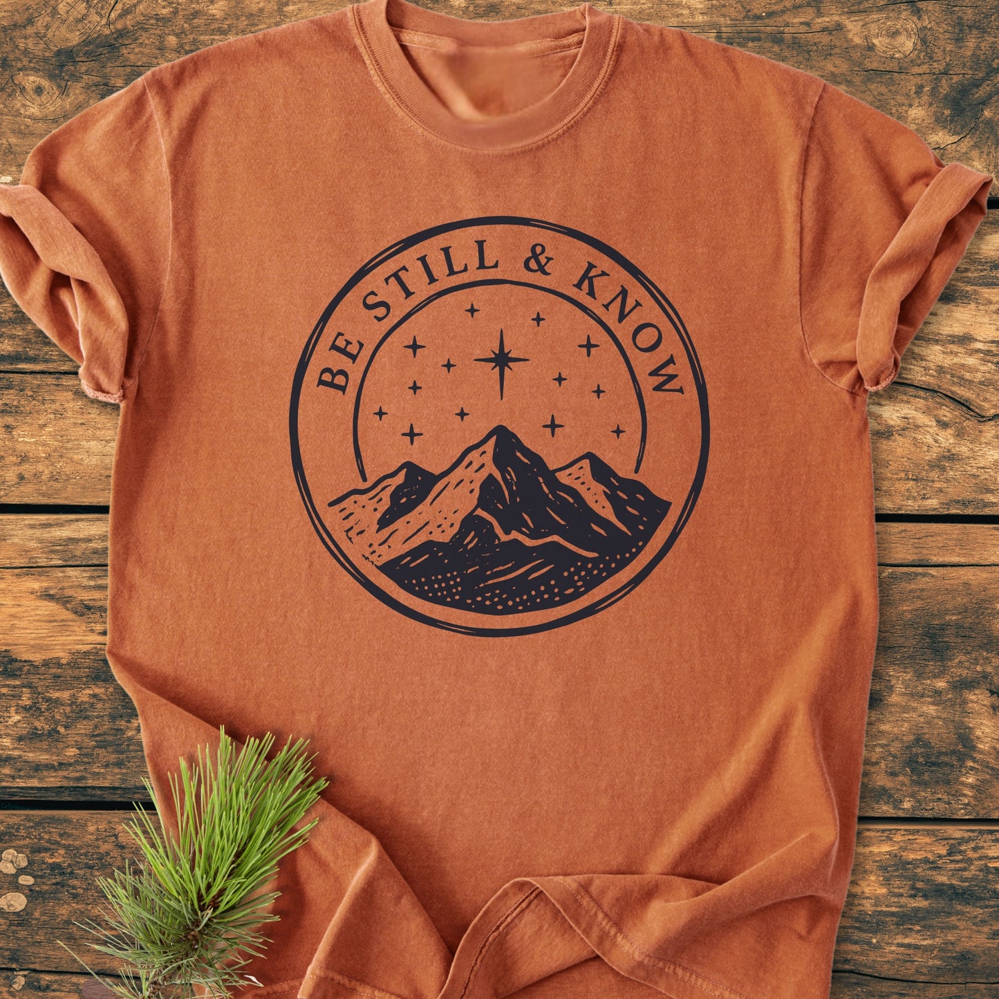 Be Still And Know, Psalm 46:10 - Vintage Wash Tee in burnt-orange, features a navy-blue mountain graphic under a starry sky, laid on a wooden surface with a pine sprig accent.