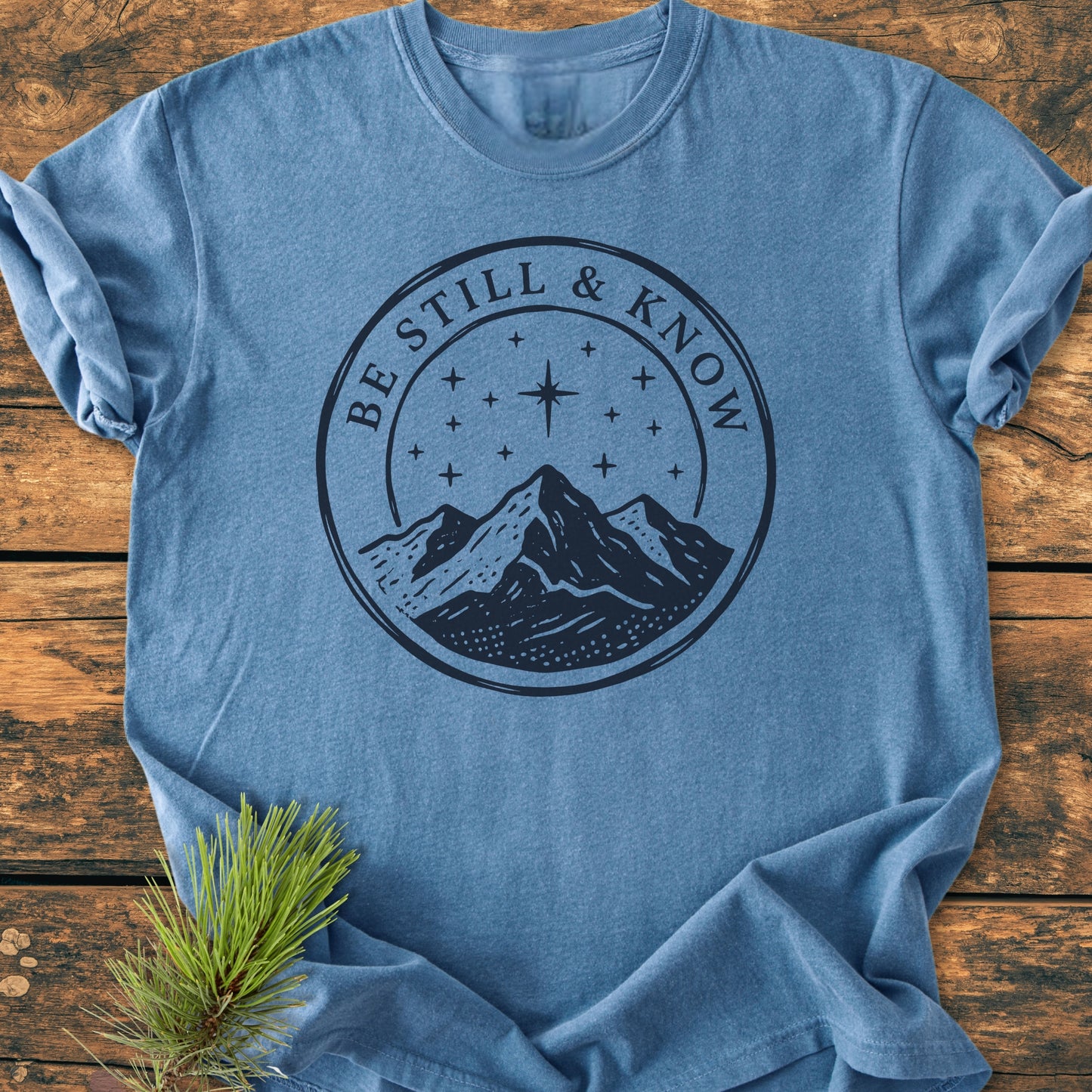 Be Still And Know, Psalm 46:10 - Vintage Wash Tee features a mountain graphic under stars on a light-blue short-sleeve, laid flat with pine branch styling on rustic wood.