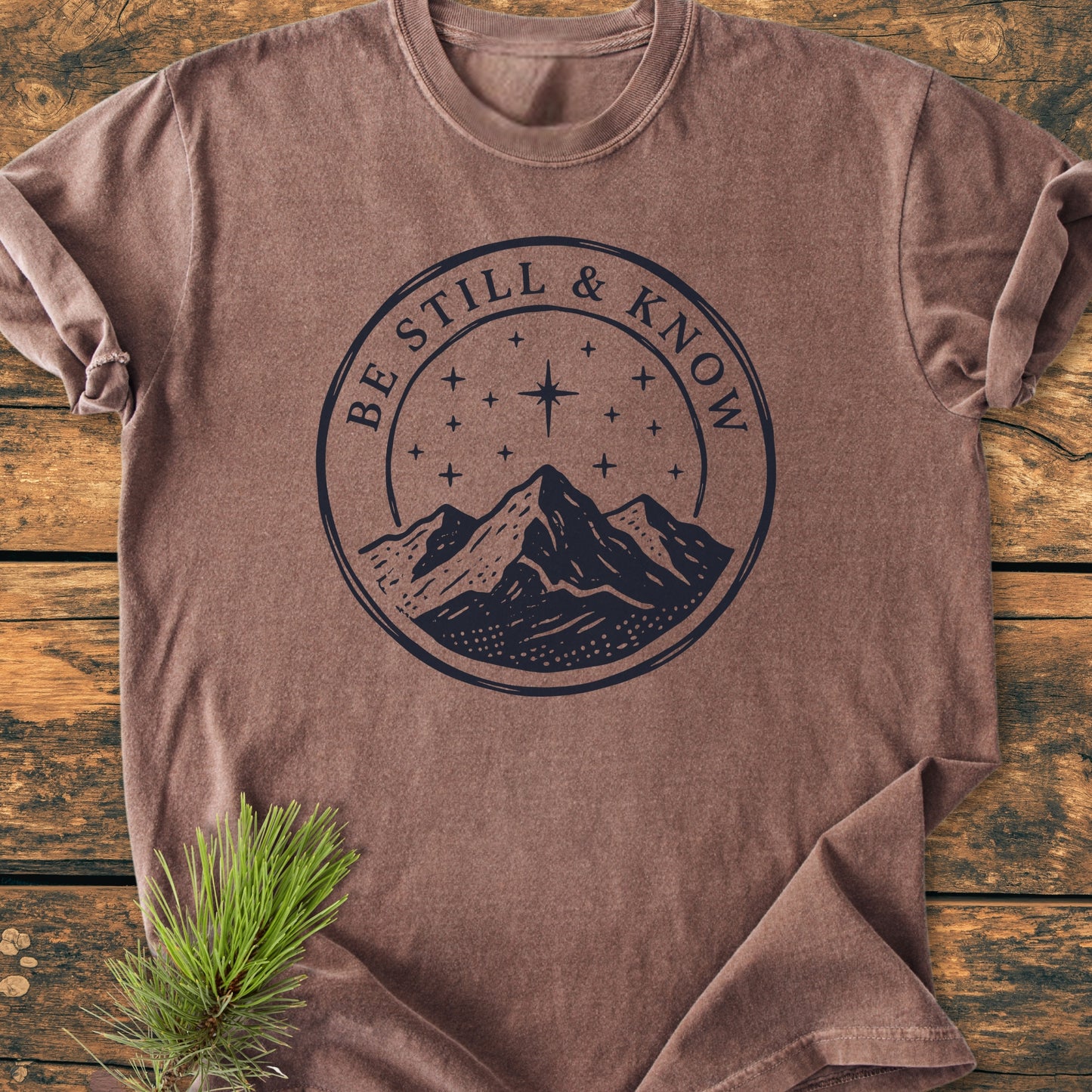 Be Still And Know, Psalm 46:10 - Vintage Wash Tee features a navy mountain graphic on a heathered-brown, folded T-shirt, resting on wood with a pine branch accent.