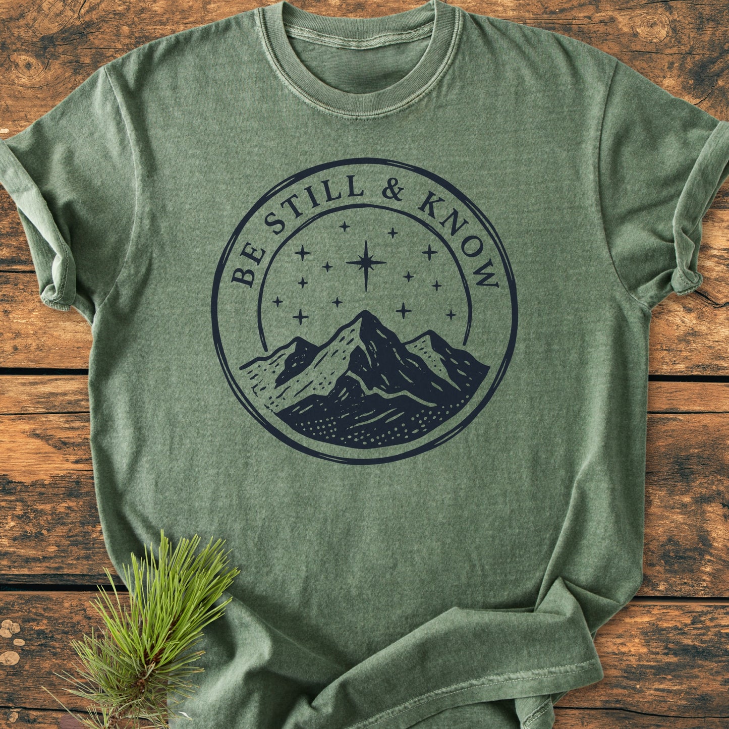 Be Still And Know, Psalm 46:10 - Vintage Wash Tee with mountain emblem and starry sky, folded on wood. Includes pine bough accent, emphasizing nature-inspired design in soft, garment-dyed cotton.