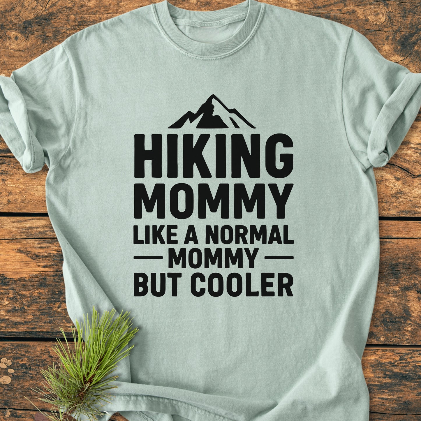 Hiking Mommy - Vintage Heavyweight Tee featuring a mountain silhouette and bold text. Displayed on a wooden surface, showcasing the relaxed fit and durable stitching of this soft, garment-dyed T-shirt.