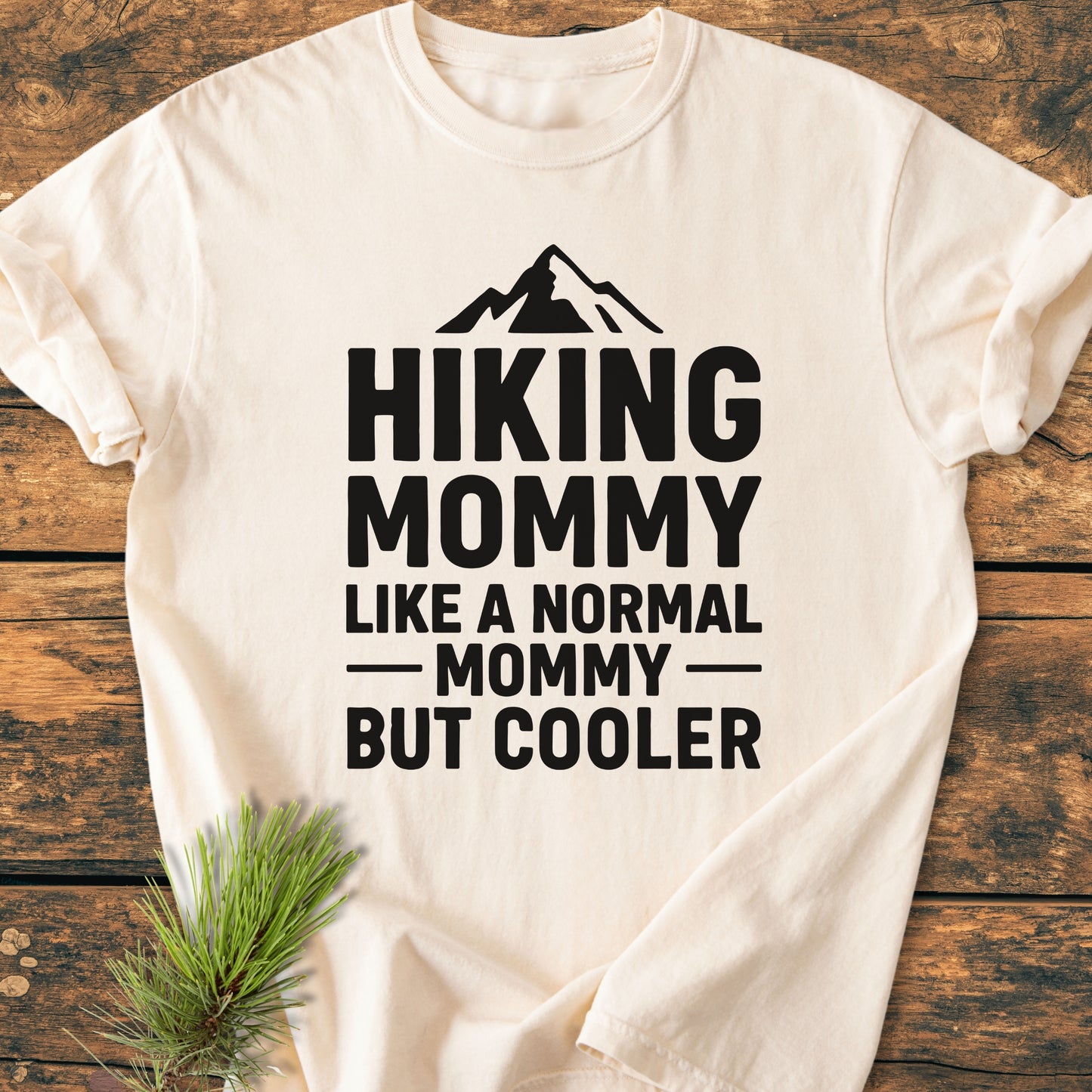 Hiking Mommy - Vintage Heavyweight Tee with bold lettering and mountain graphic, laid flat on wood. Features durable stitching and soft-washed cotton for comfort. Pine sprig accents the presentation.