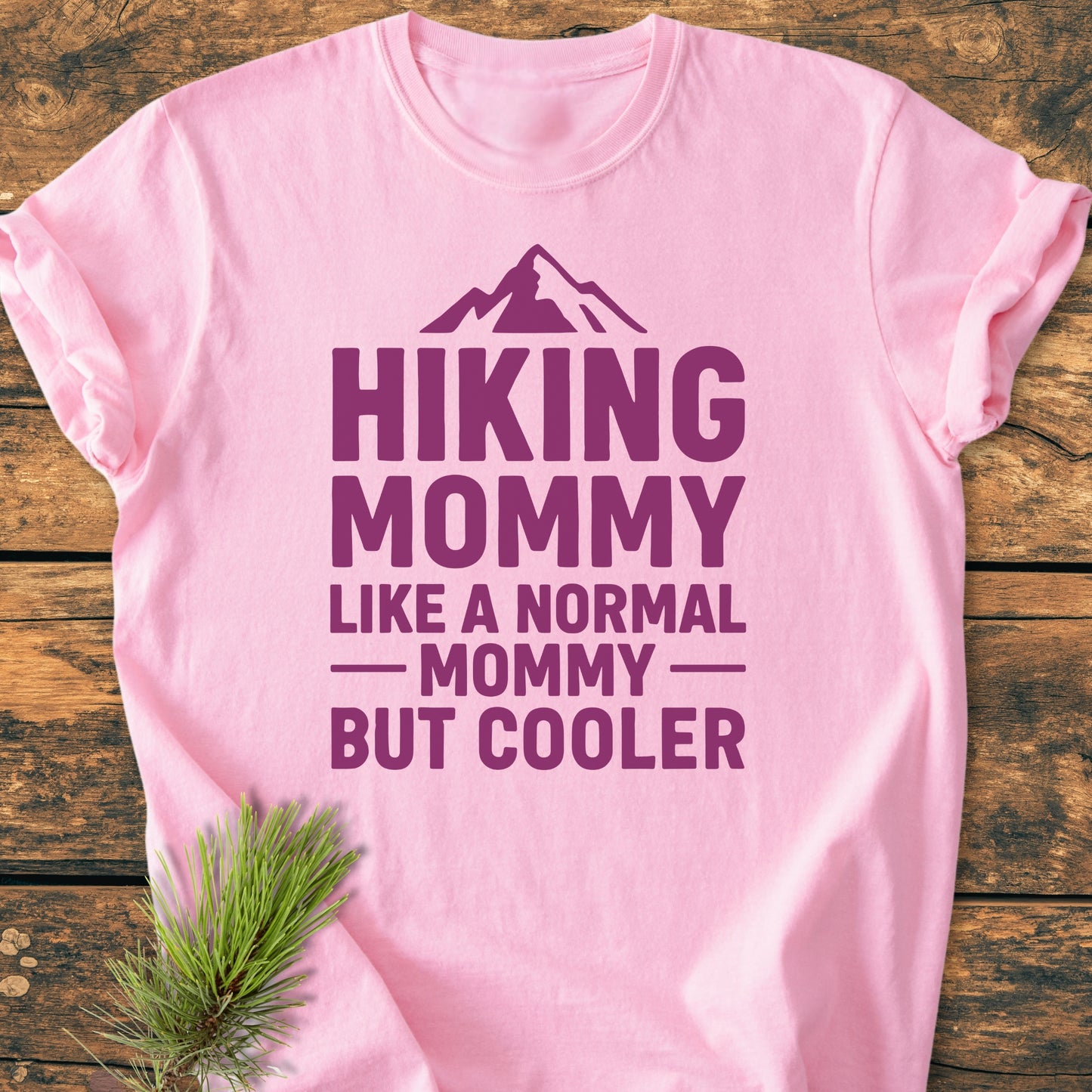 Light pink Hiking Mommy - Vintage Heavyweight Tee with mountain graphic and bold text, laid on wood. Pine twig accentuates the soft, durable, relaxed-fit design perfect for everyday wear.