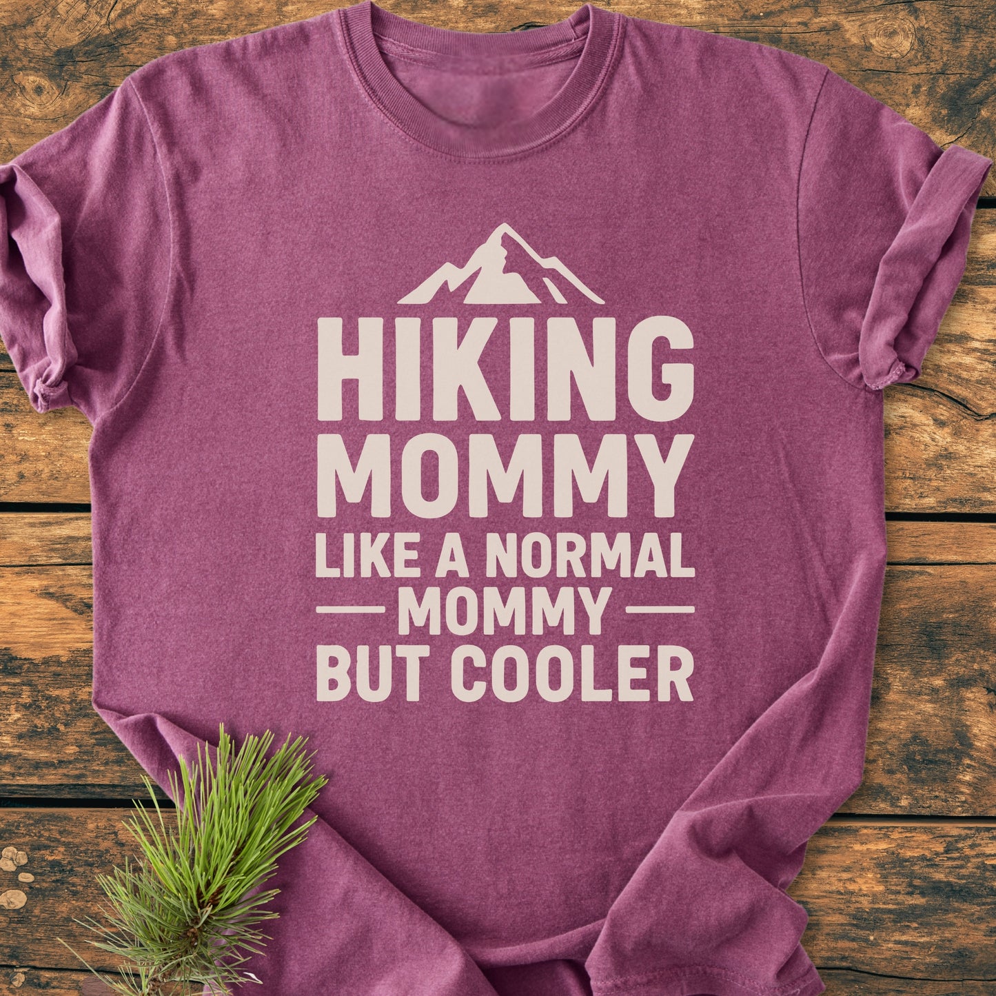 Hiking Mommy - Vintage Heavyweight Tee: A purple/maroon crew-neck T-shirt with HIKING MOMMY LIKE A NORMAL MOMMY — BUT COOLER printed in cream-white, styled with a pine sprig on rustic wood.