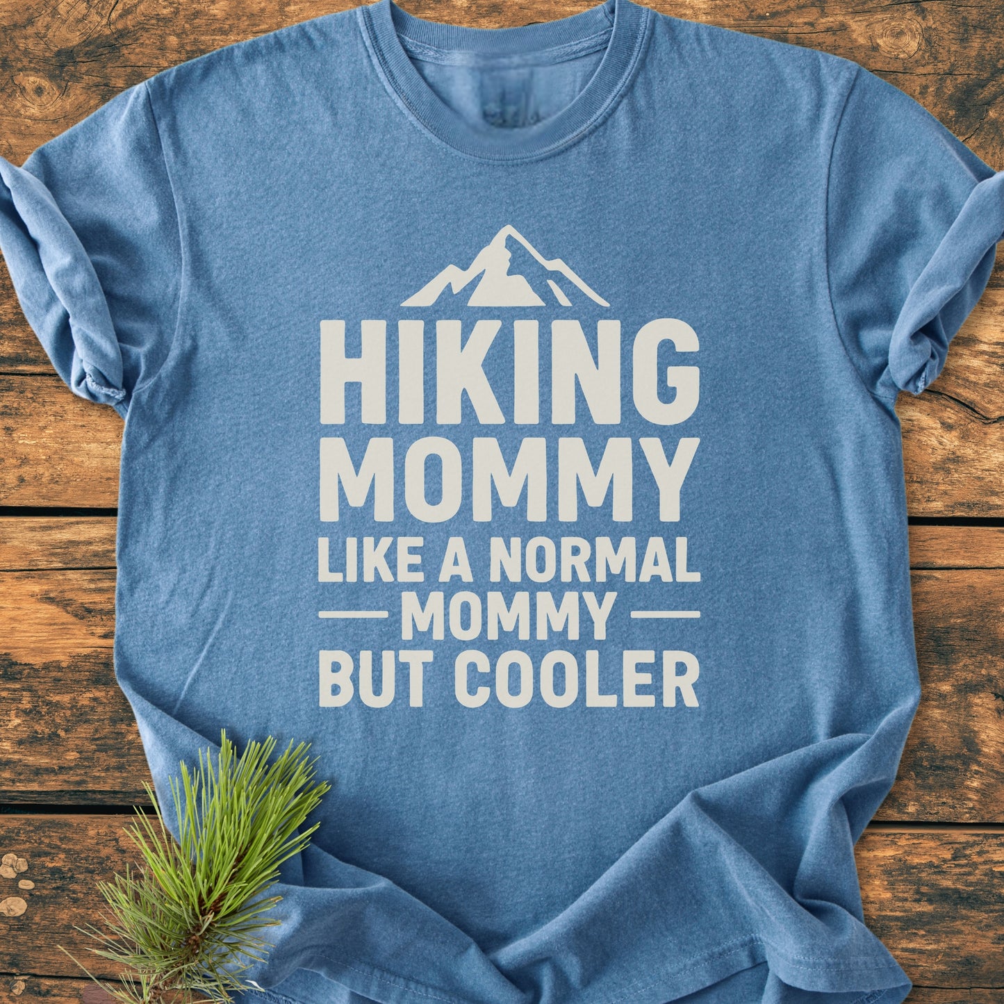 Heather-blue Hiking Mommy - Vintage Heavyweight Tee with a mountain graphic and slogan, laid flat on wood. Pine branch accentuates the cozy, relaxed fit and durable design of this ring-spun cotton tee.