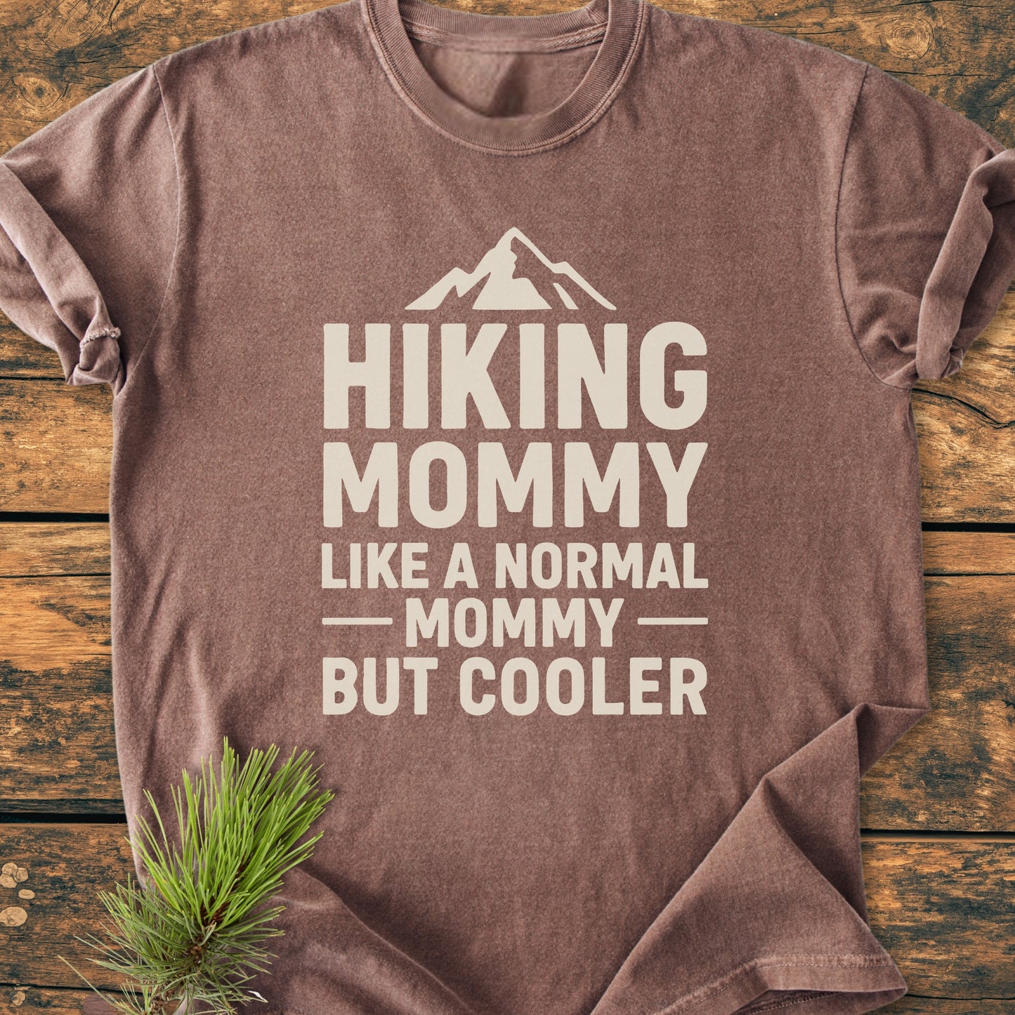 The Hiking Mommy - Vintage Heavyweight Tee features bold off-white text and a mountain icon on a brown shirt, laid flat on wood, with a pine sprig accentuating its outdoor theme.