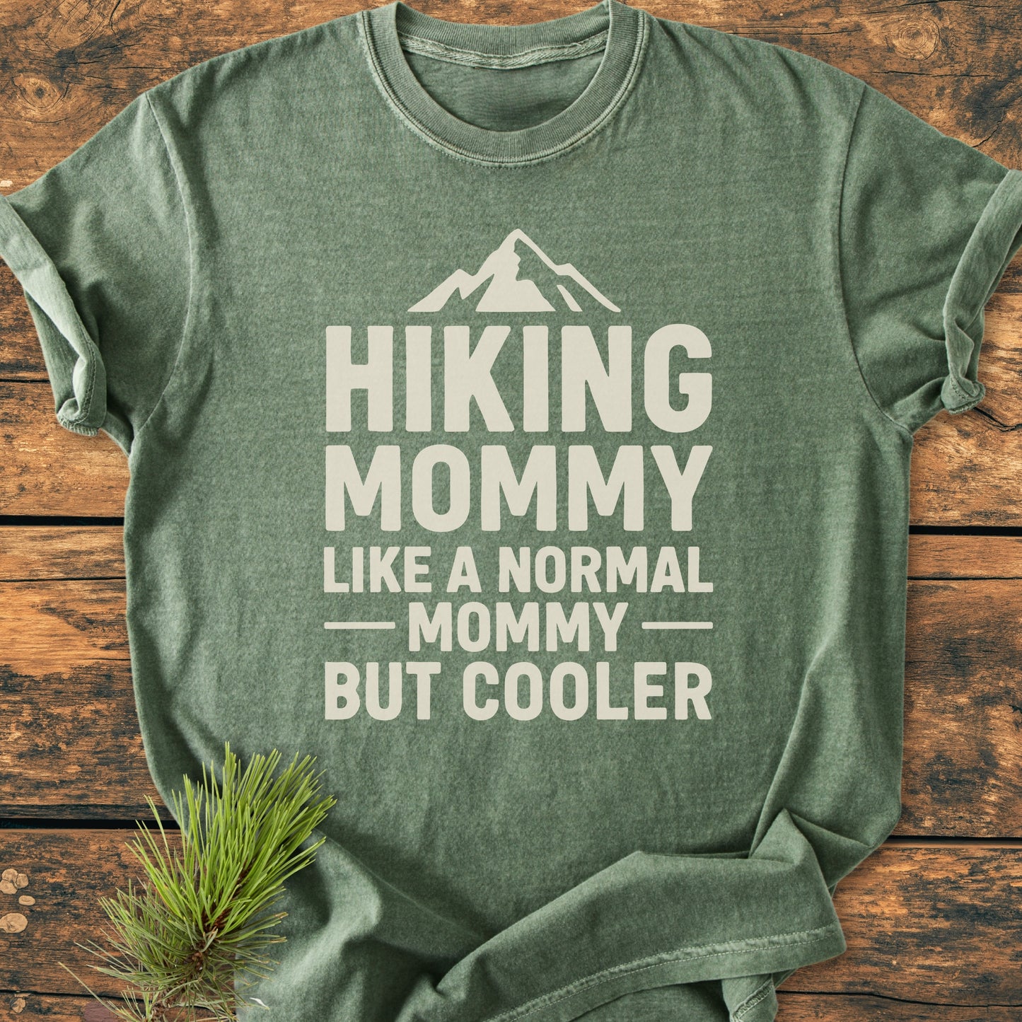 Sage-green Hiking Mommy - Vintage Heavyweight Tee with off-white text and mountain icon, laid on wood; pine sprig beside, showcasing its outdoorsy theme and relaxed, durable design.