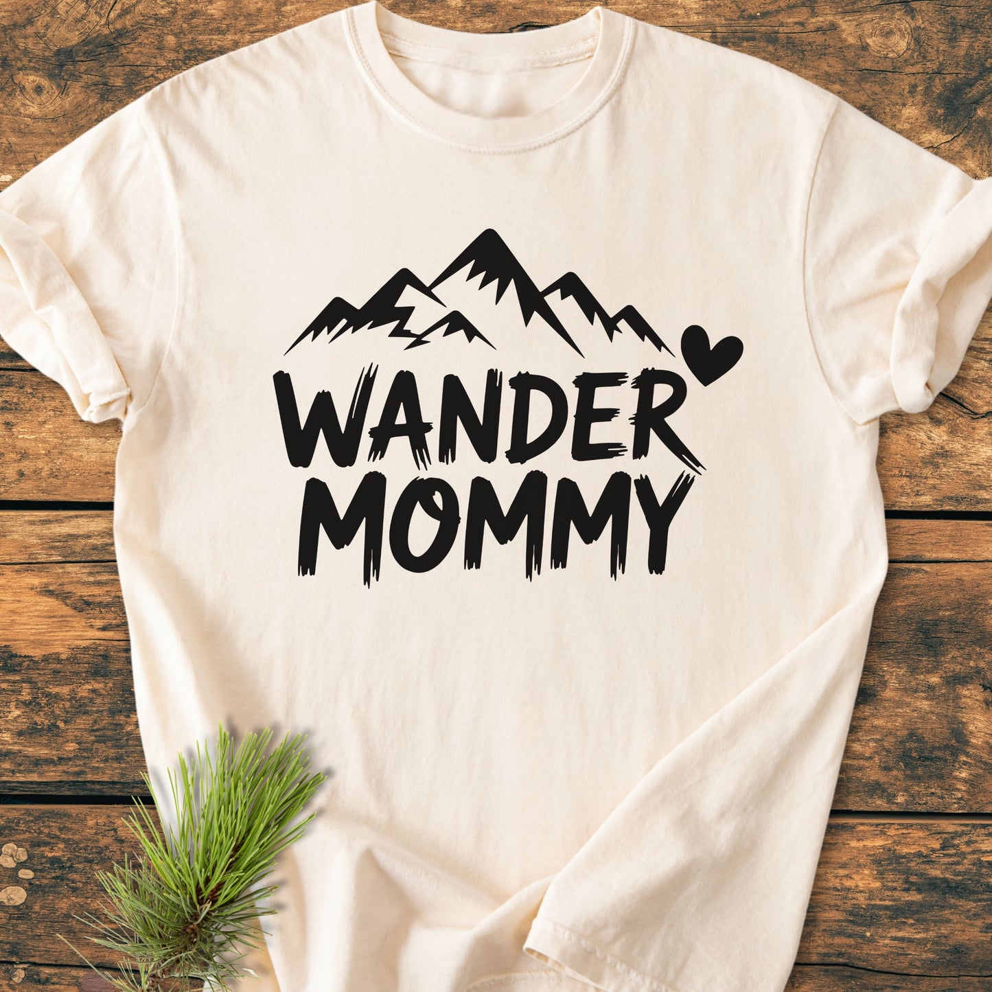 Wander Mommy - Vintage Heavyweight Tee features a mountain graphic with text on a cream-colored T-shirt, laid flat on wood, paired with a pine sprig for a natural touch.