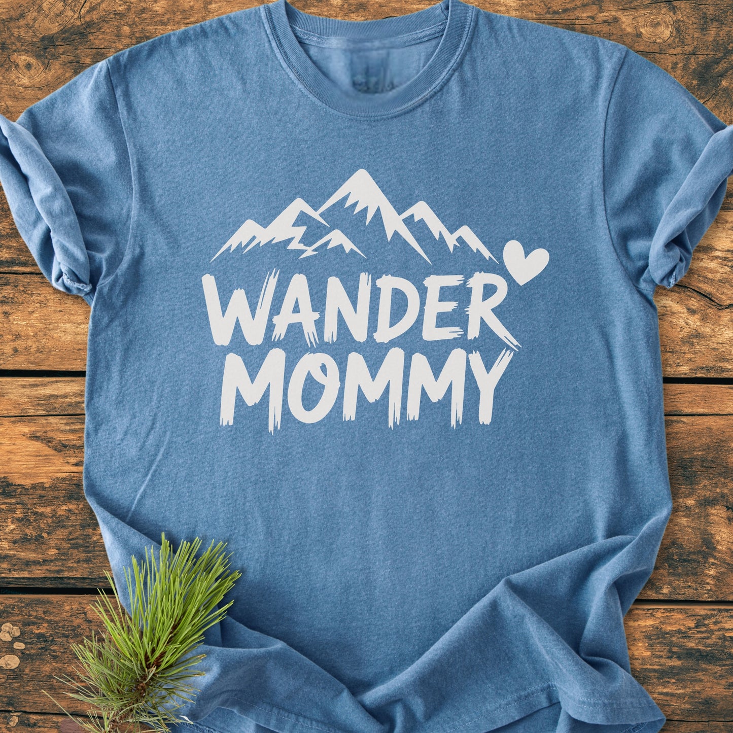 Flat-lay of the Wander Mommy - Vintage Heavyweight Tee with a white mountain graphic on a rustic surface, accompanied by a pine branch for a cozy, nature-inspired touch.
