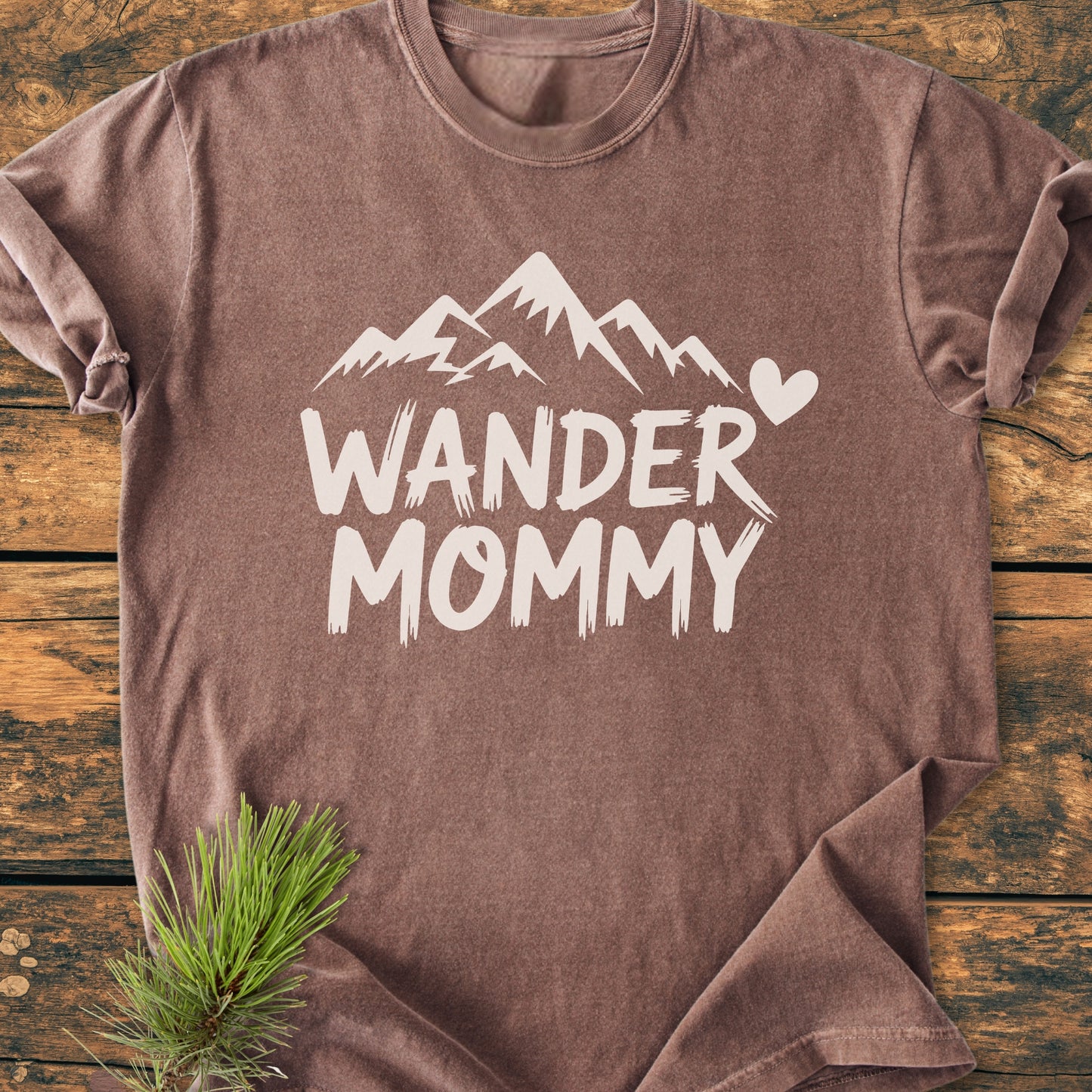 Wander Mommy - Vintage Heavyweight Tee flat-lay showing a brown short-sleeve shirt with mountain silhouette and text, styled on a wooden surface with a pine sprig for natural accent.