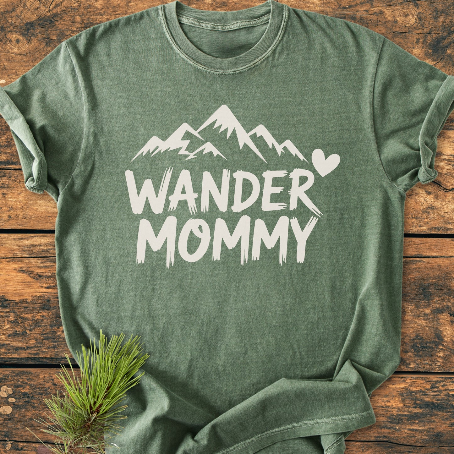 Wander Mommy - Vintage Heavyweight Tee features a hand-painted mountain design and heart on the chest, displayed flat on wood with pine needles, highlighting its relaxed fit and durability.