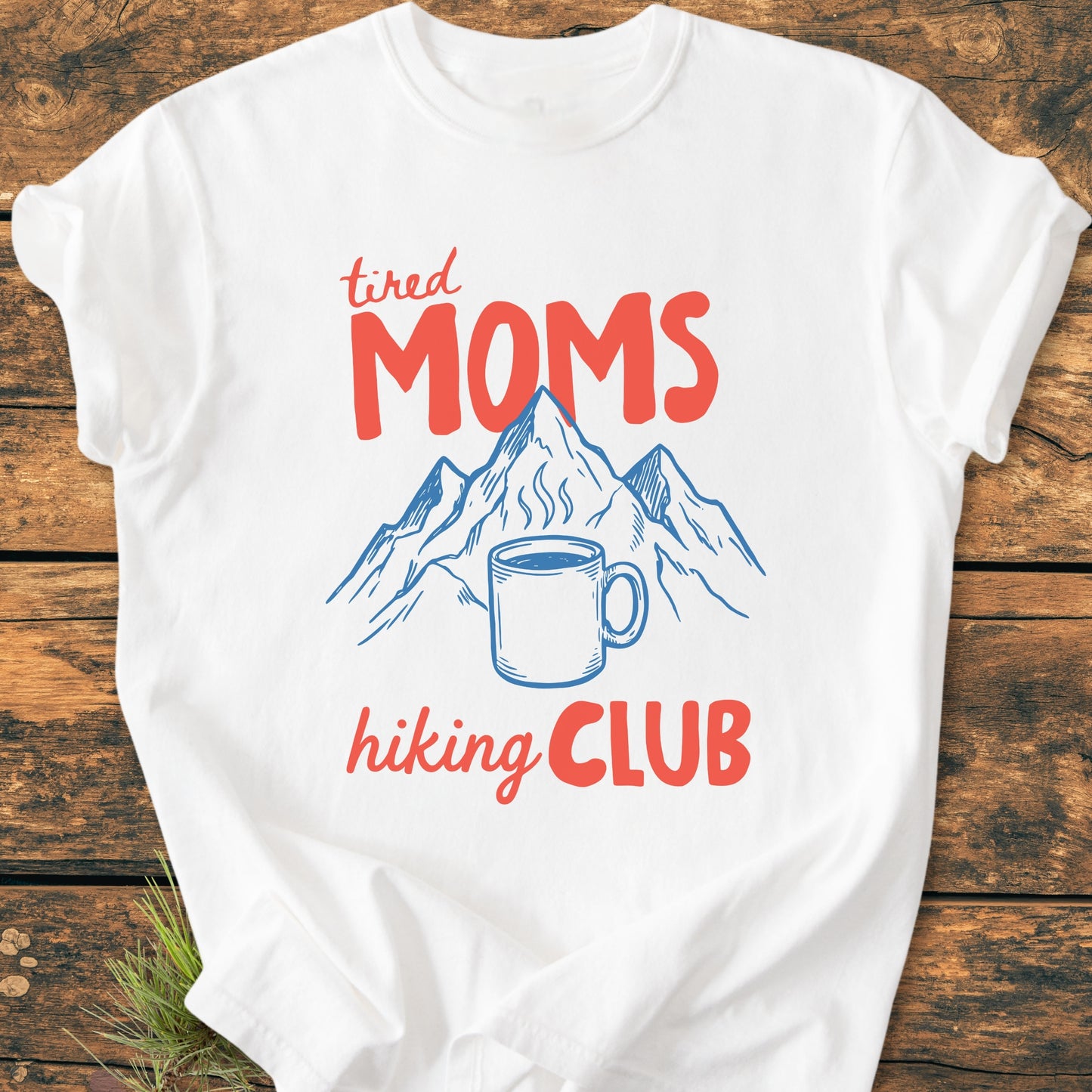 Tired Moms Hiking Club - Vintage Heavyweight Tee features a mountain line-drawing with a coffee mug. The white, relaxed-fit shirt lies on a wooden surface, highlighting its durable, soft-washed cotton fabric.