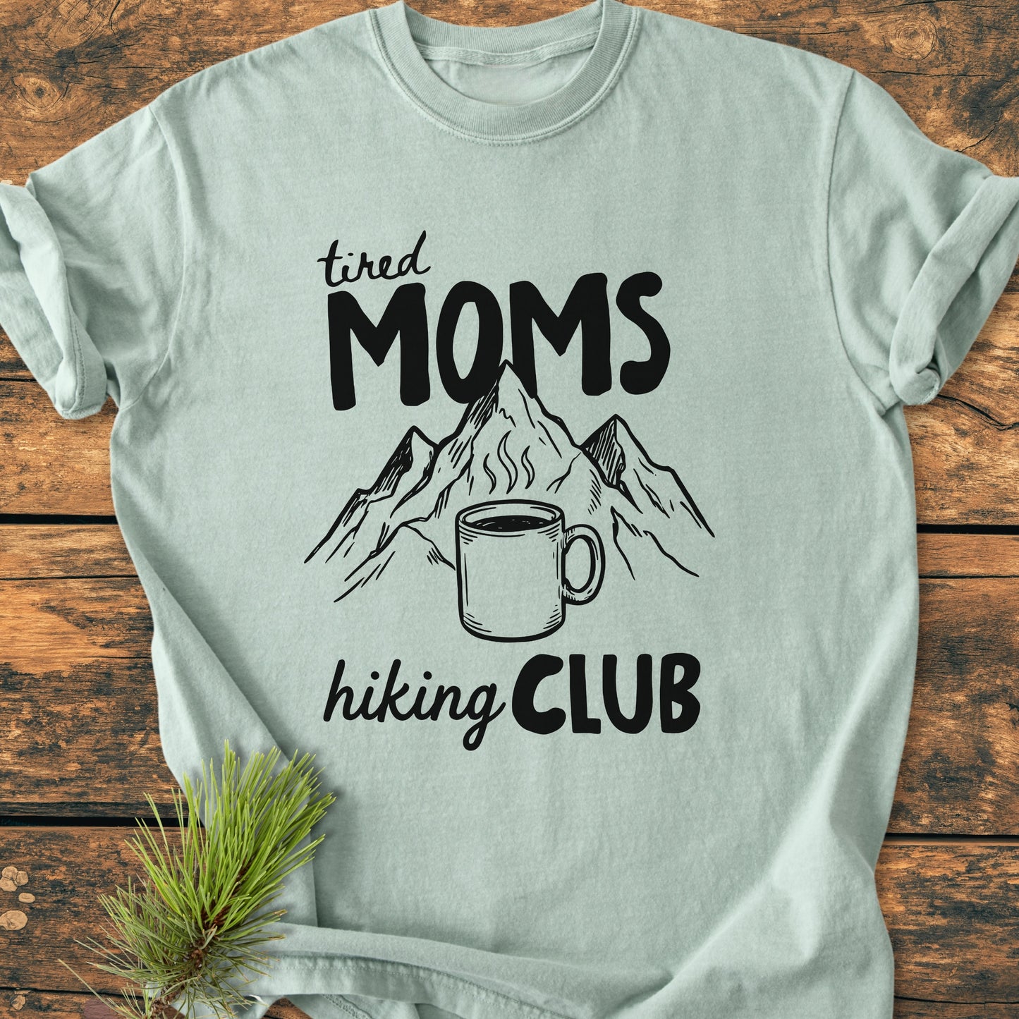 Tired Moms Hiking Club - Vintage Heavyweight Tee features a tired MOMS print with mountain peaks and coffee mug, laid on wood with pine sprig. Made from soft, durable, garment-dyed cotton.