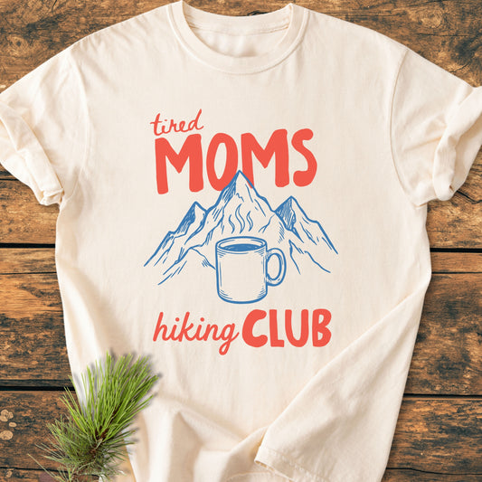 Tired Moms Hiking Club - Vintage Heavyweight Tee features bold red lettering and a mountain with coffee mug illustration, laid on wood with a pine sprig, showcasing its cozy, relaxed fit.