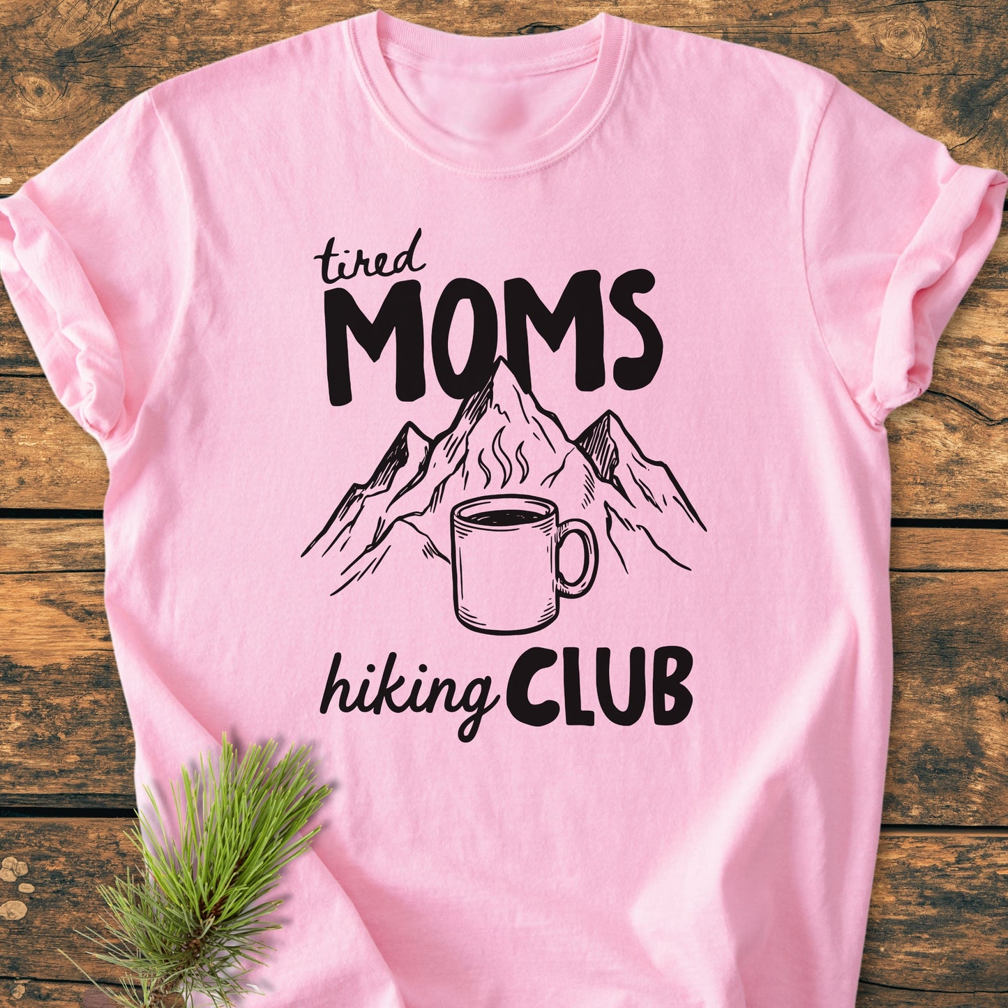 Tired Moms Hiking Club - Vintage Heavyweight Tee features bold black lettering and mountain art with a steaming mug. Presented on a wooden surface, highlighting its soft-washed, relaxed Comfort Colors design.