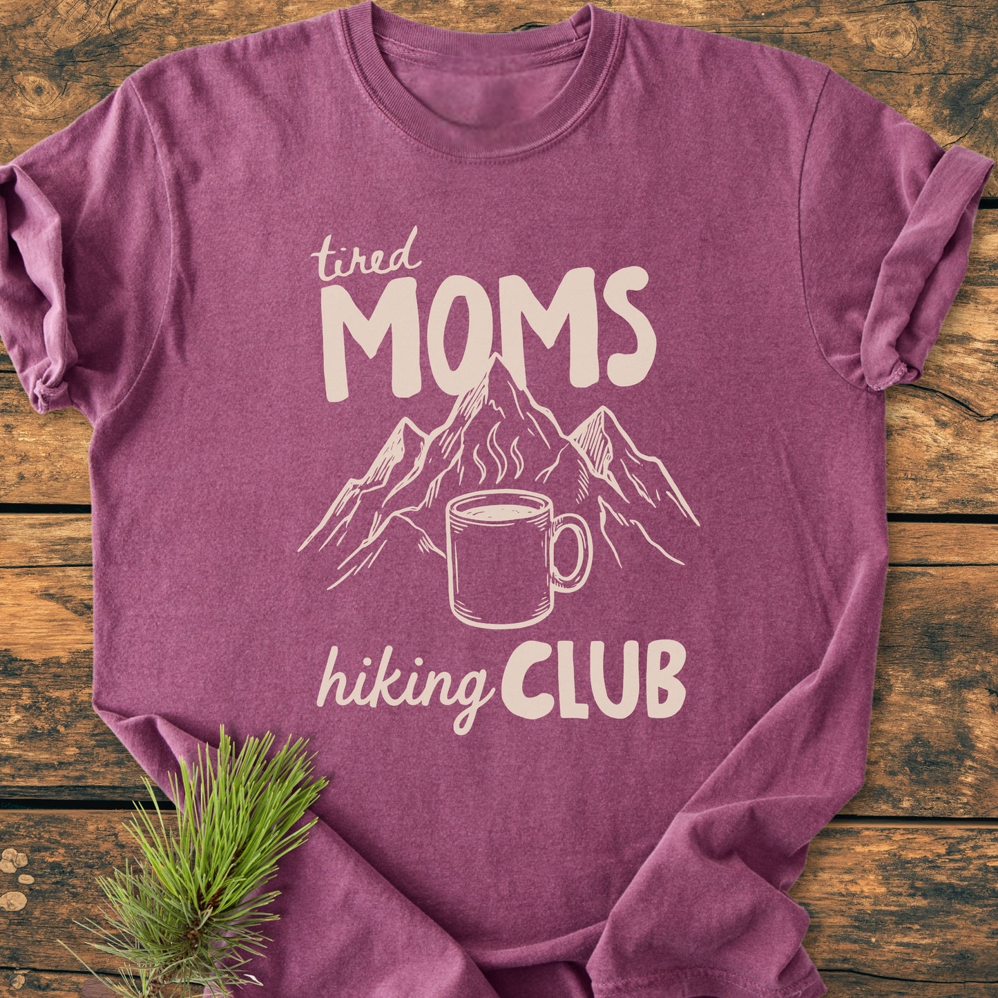 Flat-lay of the Tired Moms Hiking Club - Vintage Heavyweight Tee, featuring a hand-drawn mountain and coffee mug design, styled with a pine sprig on rustic wood, showcasing its relaxed fit.