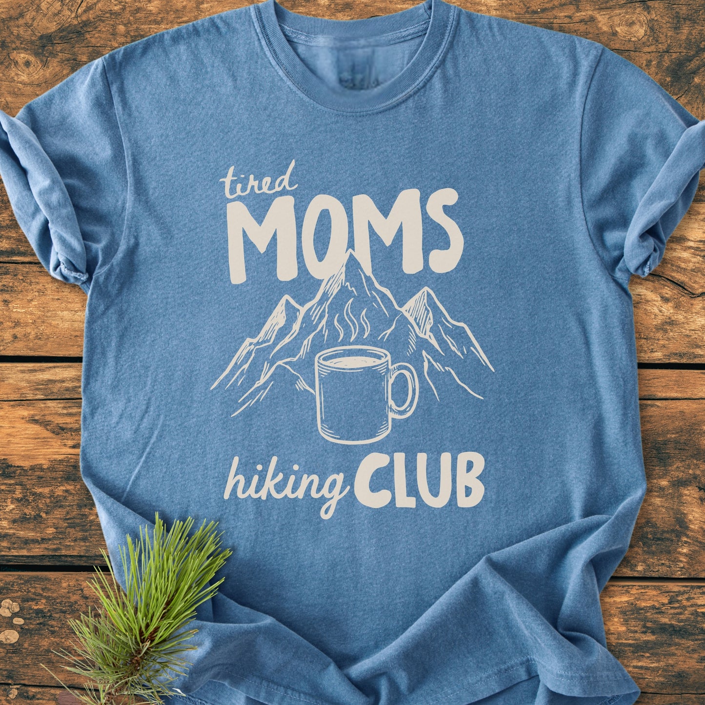 Tired Moms Hiking Club - Vintage Heavyweight Tee displays a steaming coffee mug with mountain peaks graphic, laid on a wooden surface. Crafted from soft, durable, ring-spun cotton for everyday comfort.