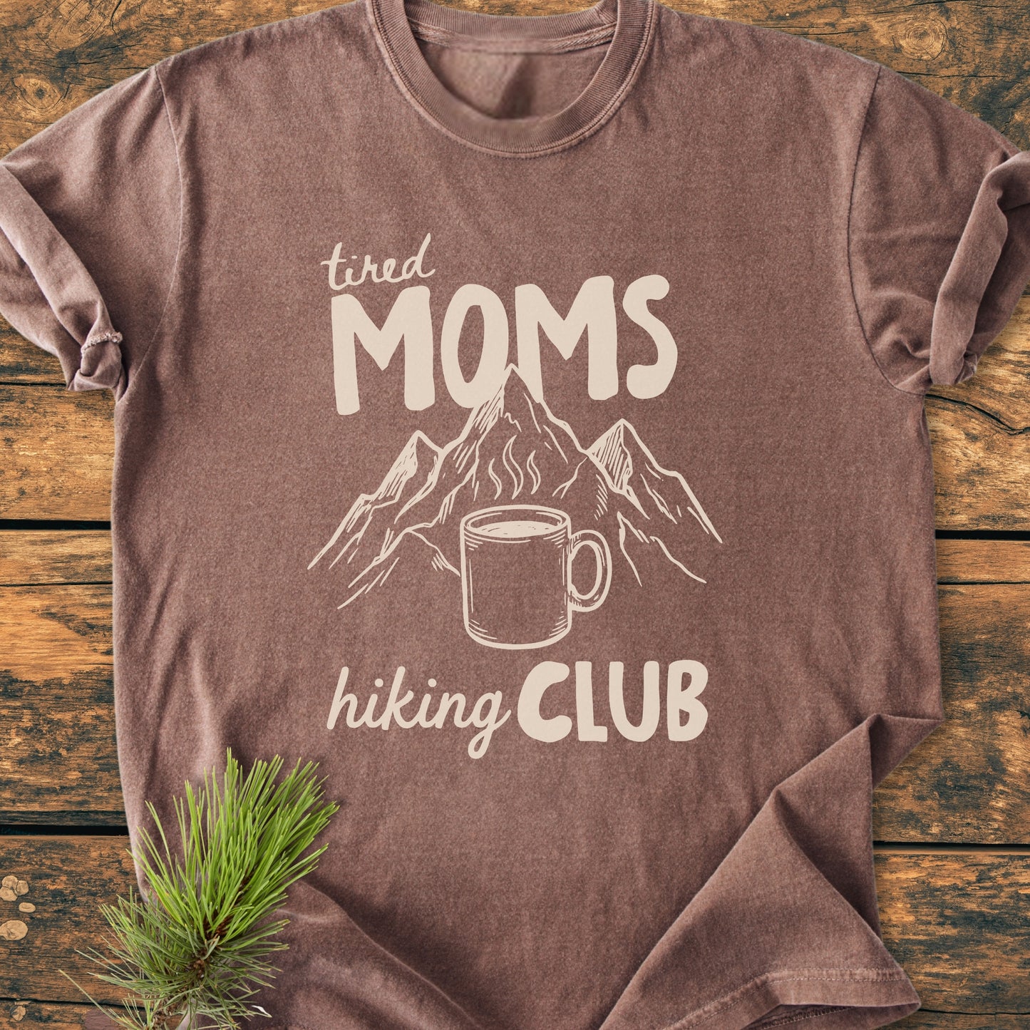 Tired Moms Hiking Club - Vintage Heavyweight Tee featuring a mountain and coffee mug design. Laid on wood, with pine accent, showcasing its relaxed fit and durable, soft-washed cotton fabric.