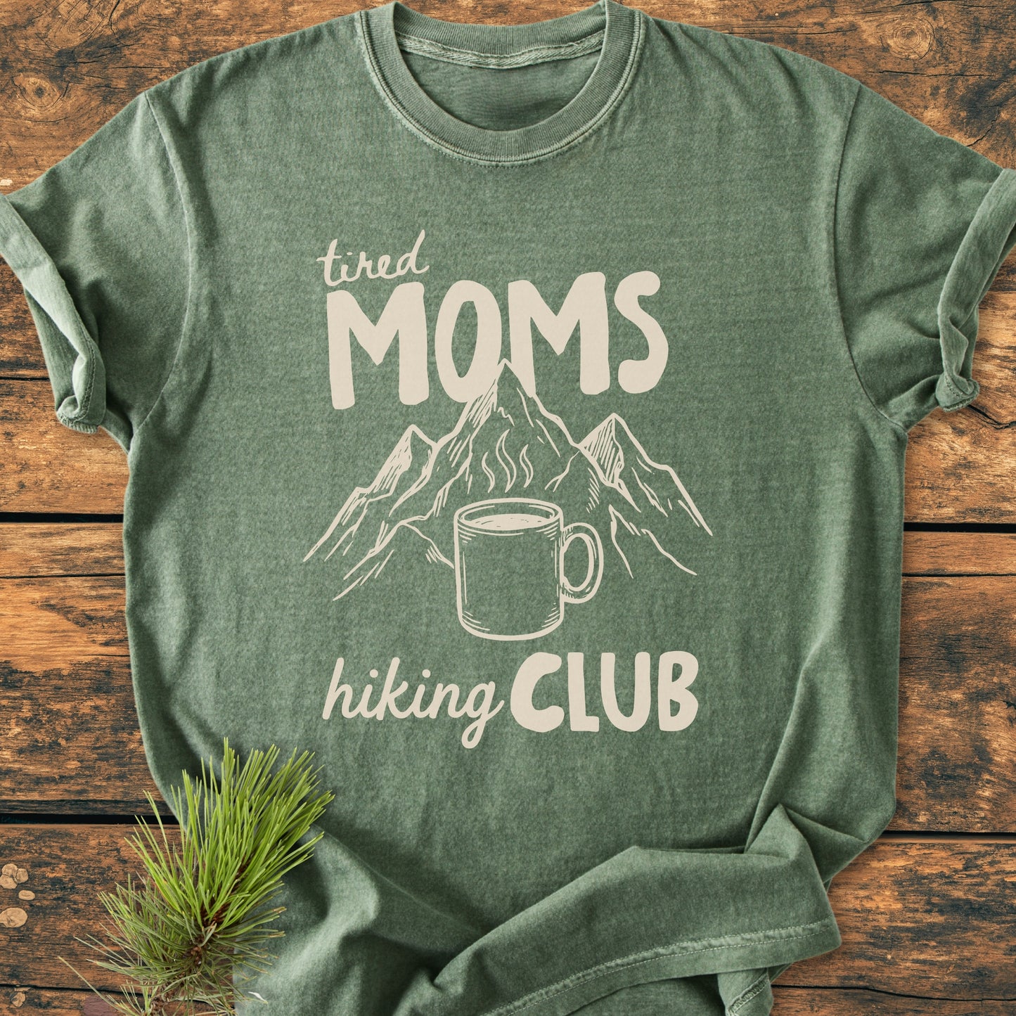 The Tired Moms Hiking Club - Vintage Heavyweight Tee features a mountain range and coffee mug graphic on a soft green heather t-shirt, laid on wood, with a pine sprig beside it.