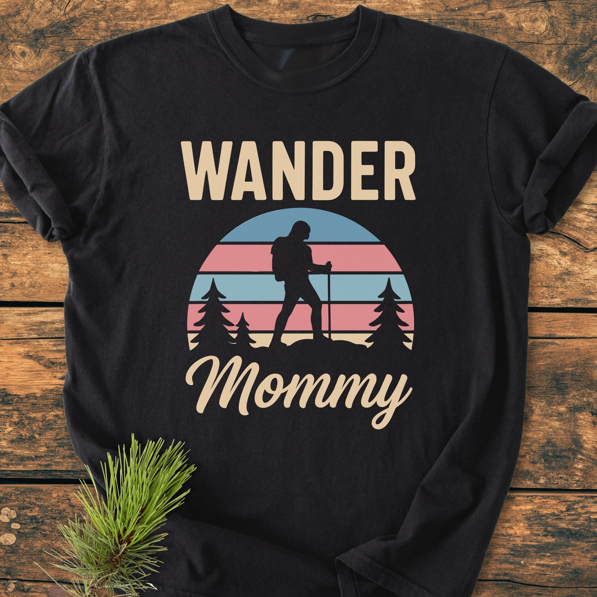 Wander Mommy Retro - Vintage Heavyweight Tee features a black tee with 'WANDER Mommy' and a retro-striped sunset graphic, showcasing a hiker silhouette, laid on a wooden surface with greenery.