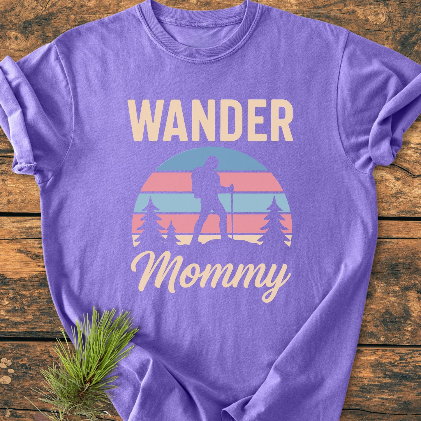 Wander Mommy Retro - Vintage Heavyweight Tee featuring a hiker graphic with trekking poles, sun backdrop, and pine trees. Laid on a wooden surface with a pine sprig nearby.