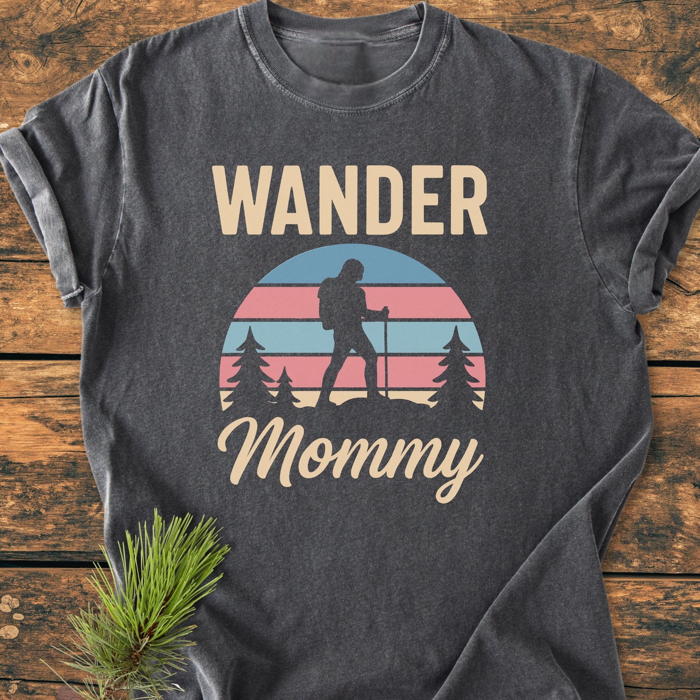 Heather-gray Wander Mommy Retro Tee featuring a graphic of a hiker among pine trees and a striped sun, laid on wood. Highlighting its relaxed fit, soft-washed texture, and durable build.