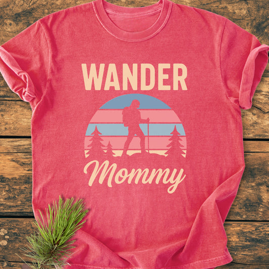 Wander Mommy Retro - Vintage Heavyweight Tee features a hiker graphic with sunset and pine trees on a heather-red short-sleeve. Laid flat on wood, showcasing its relaxed, soft-washed fabric.