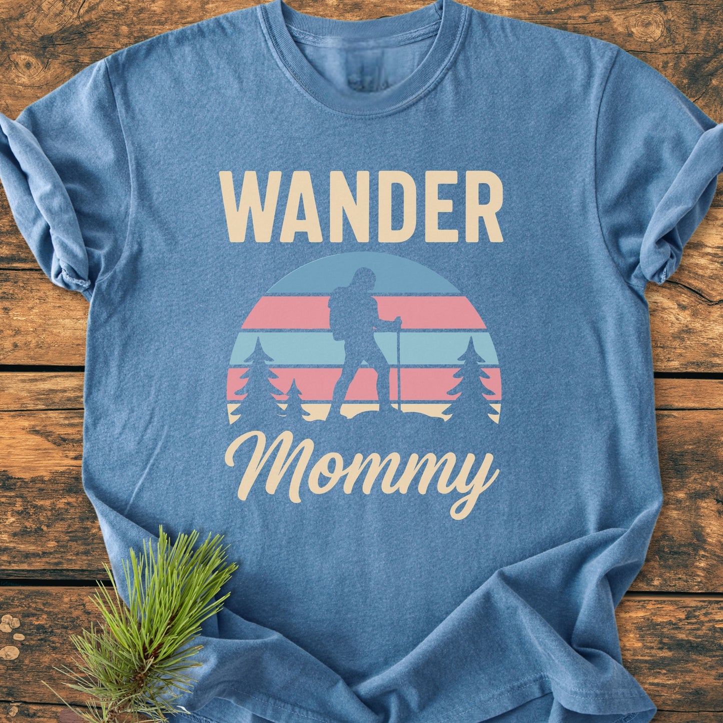 The Wander Mommy Retro - Vintage Heavyweight Tee in heather-blue, featuring a backpacking silhouette graphic and cream lettering, displayed on a wooden surface with a pine sprig accent.