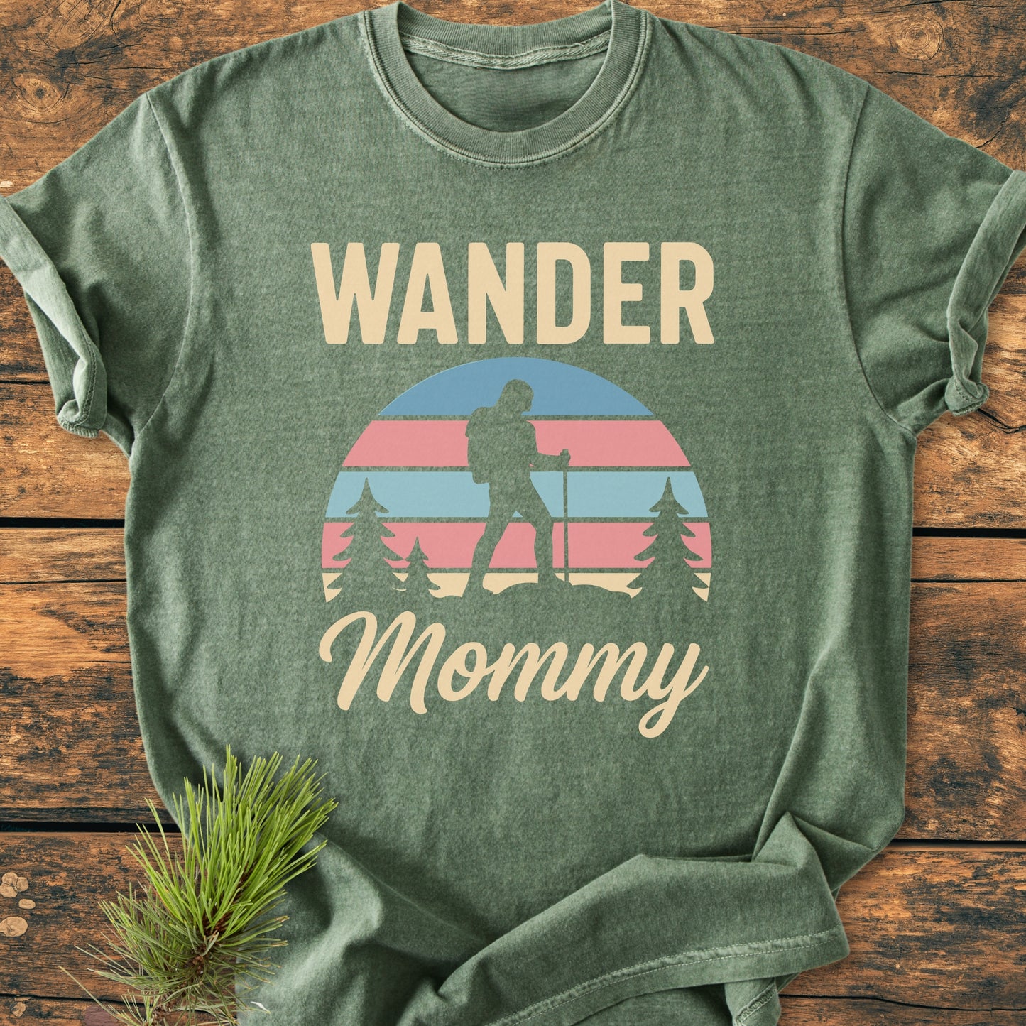 Heather-green Wander Mommy Retro - Vintage Heavyweight Tee featuring cream text and a pastel-striped hiker graphic. Laid on wood with pine sprig, showcasing its relaxed, durable design in ring-spun cotton.