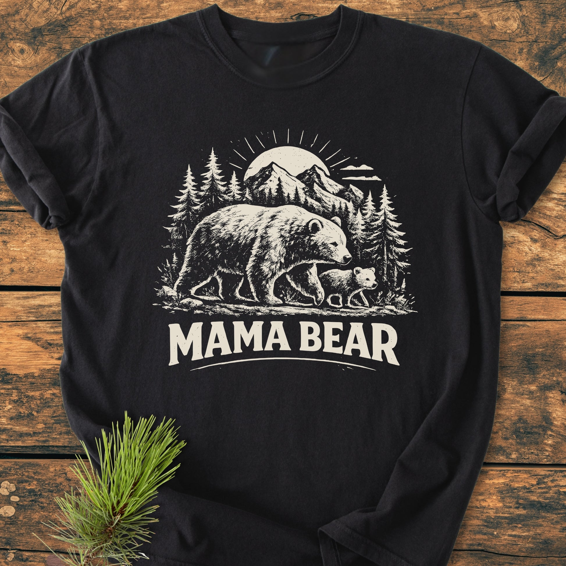 Mama Bear - Vintage Heavyweight Tee: Black T-shirt with cream graphic of a mother bear and cub in a pine forest, featuring MAMA BEAR text, displayed on a wooden surface with pine sprig.