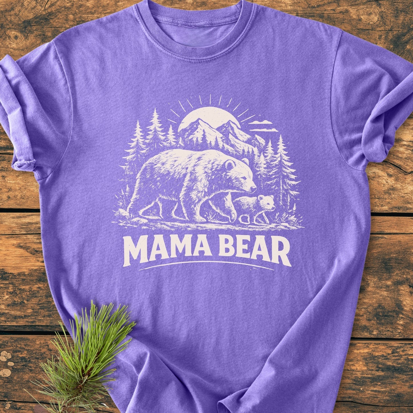 Mama Bear - Vintage Heavyweight Tee featuring a white illustration of bears and nature on a purple T-shirt, displayed on wood with a pine branch beside it, showcasing relaxed fit and durable stitching.