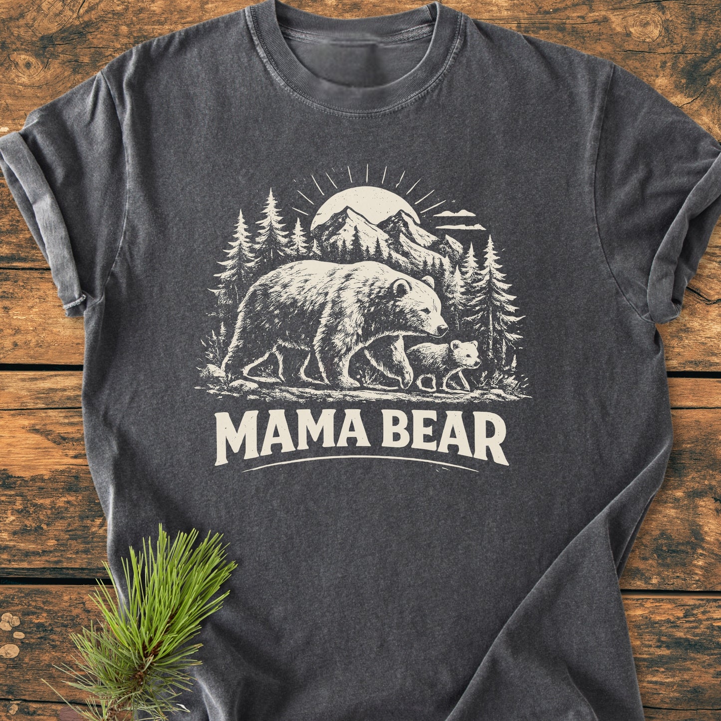 Mama Bear - Vintage Heavyweight Tee: Charcoal T-shirt with a nature scene of bears, pine trees, and mountains, printed in off-white. Features MAMA BEAR text, laid on a wooden surface with a pine sprig.