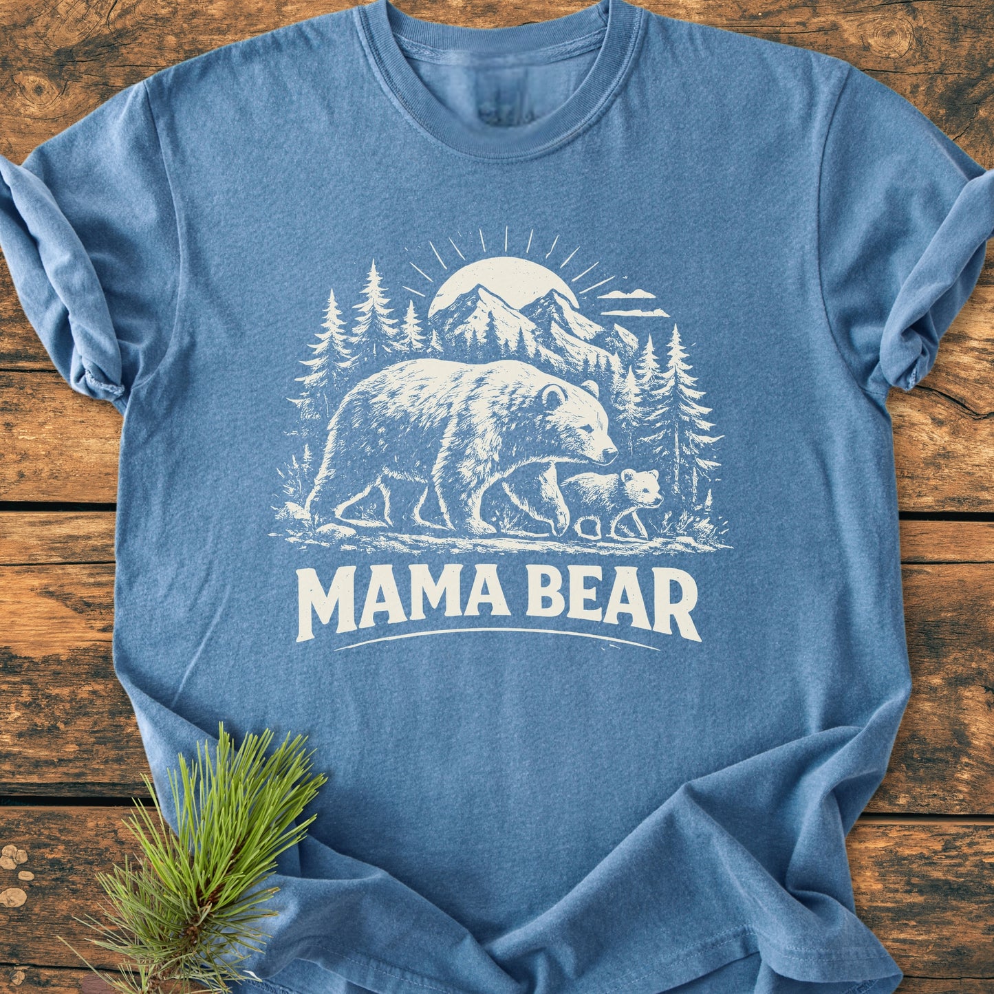 Mama Bear - Vintage Heavyweight Tee featuring a bear and cub graphic in a pine forest with mountains. Laid on rustic wood with a pine sprig, showcasing its relaxed fit and durability.