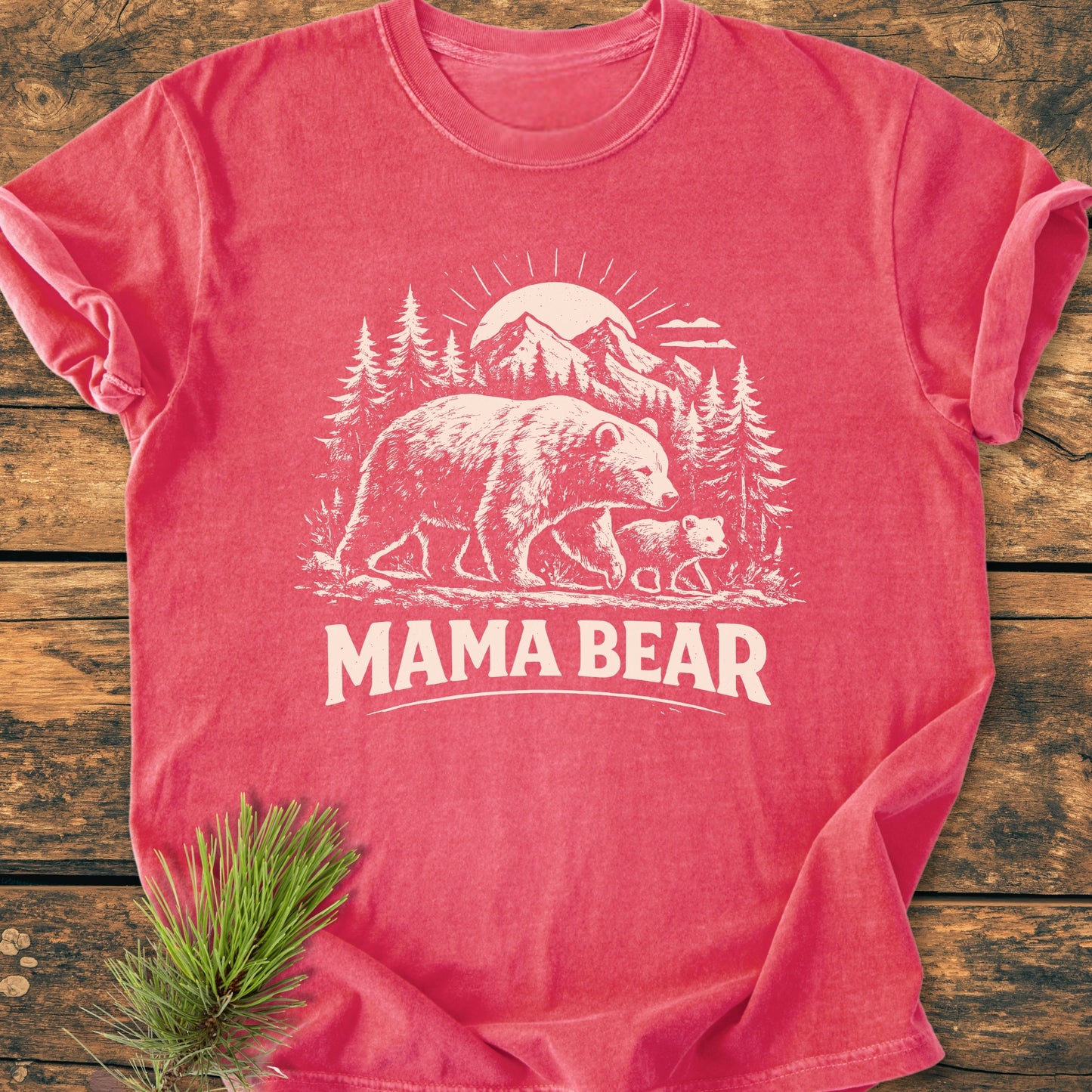 Mama Bear - Vintage Heavyweight Tee features a white forest scene with a bear, cub, and mountains, printed on a reddish-pink T-shirt, lying on wood, with a pine sprig beside it.