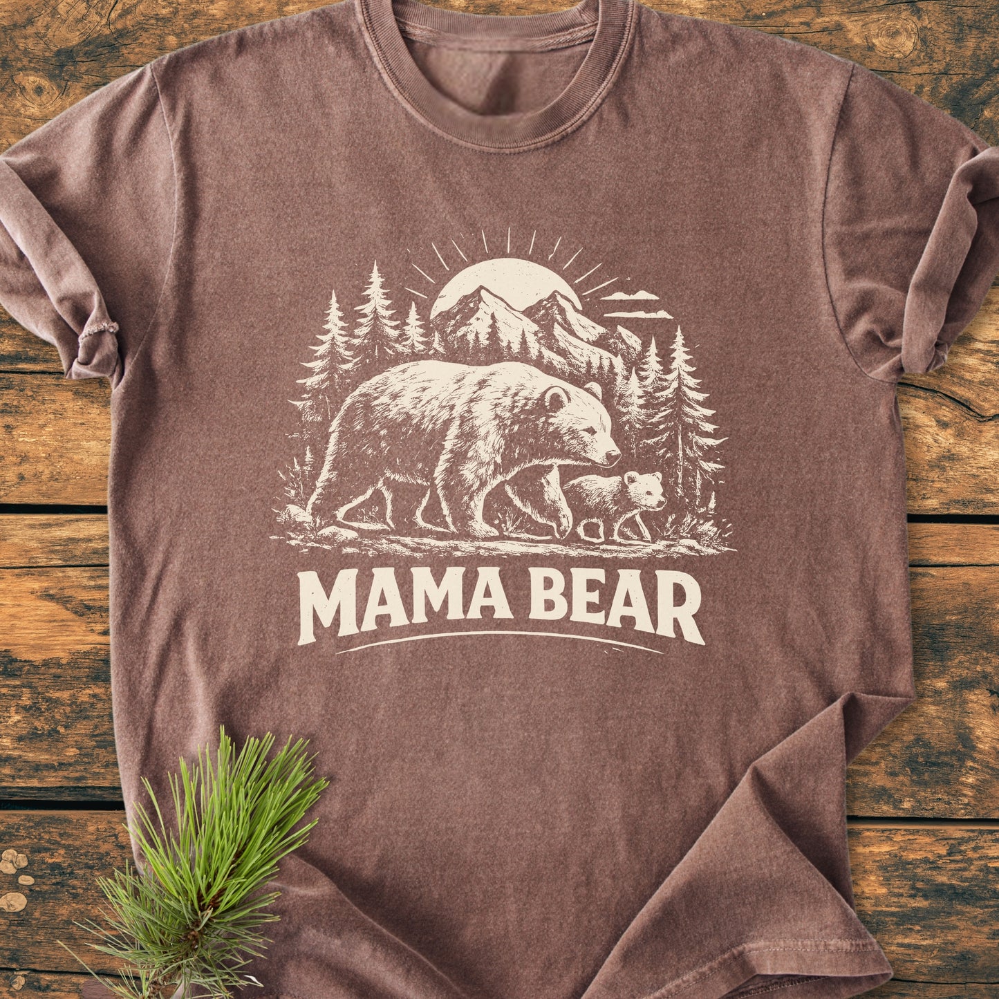 Mama Bear - Vintage Heavyweight Tee features a bear and cub print with pine trees and mountains, laid flat on wood with a pine sprig beside it. Ideal for comfort and durability.